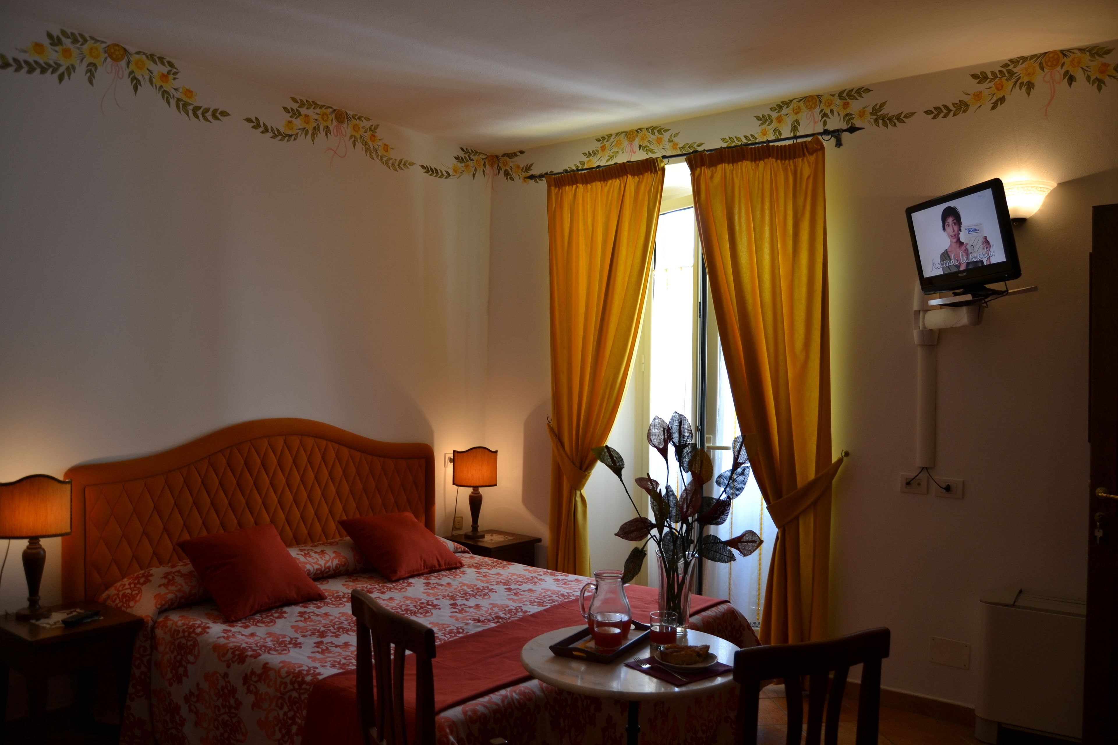 A hotel room with a bed, a table with a pitcher and a vase, a TV on the wall, and curtains.