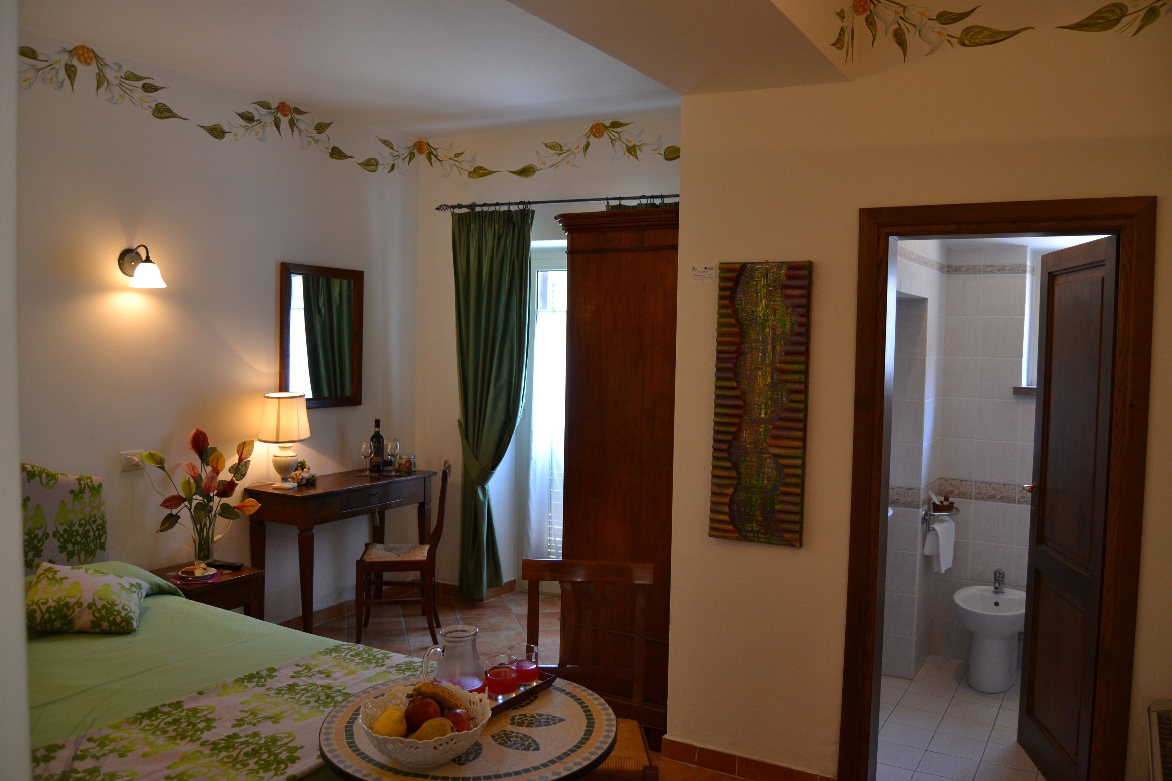 Une chambre d’hôtel avec un lit, un bureau, une chaise, une table à manger agrémentée d’un panier de fruits, une lampe et une salle de bain visible à travers une porte ouverte.