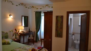 Hotelkamer met een bed, een bureau, een stoel, een eettafel met een mand fruit, een lamp en een badkamer zichtbaar door een geopende deur.