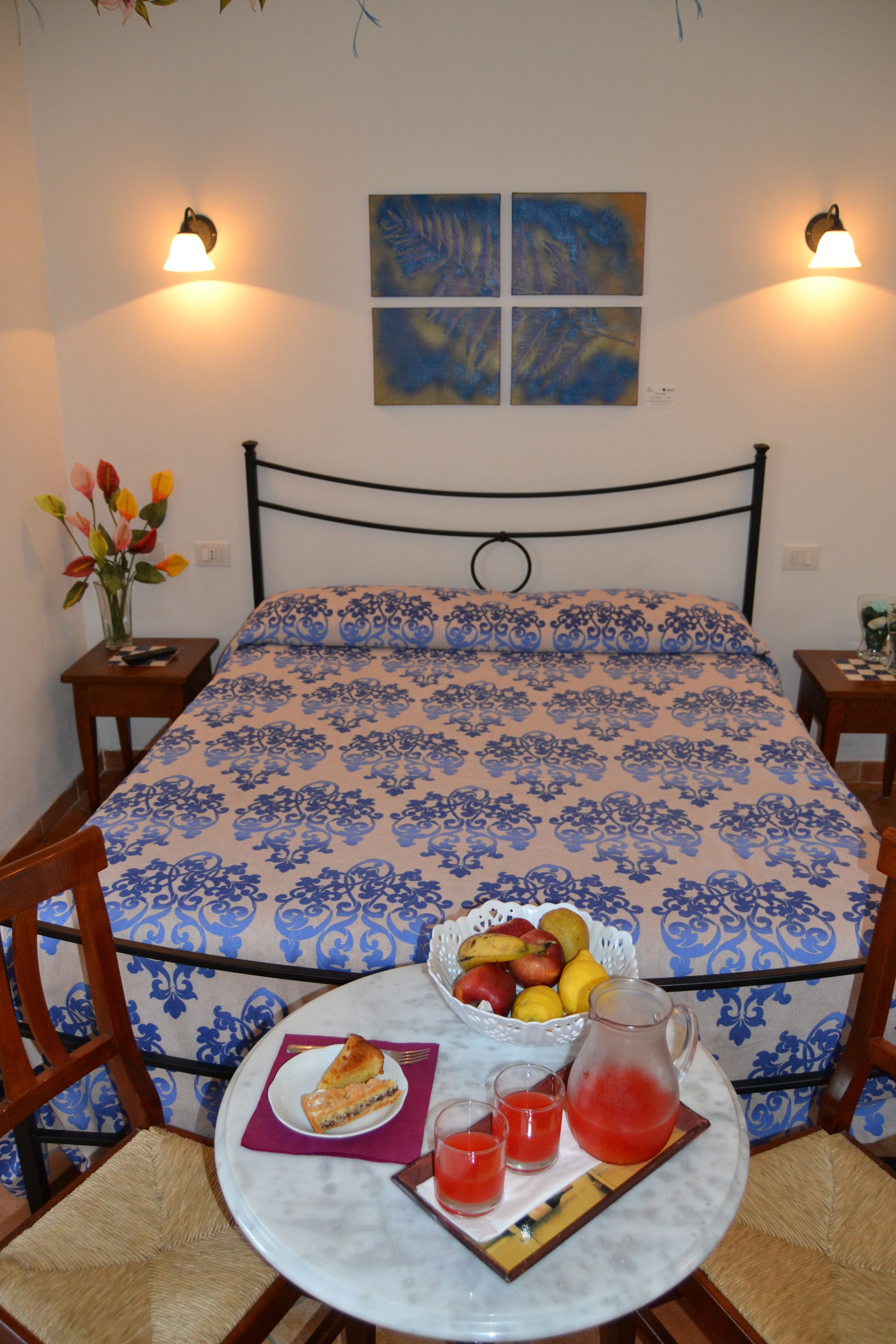 Una cama bien tendida con una colcha azul estampada con flores, una mesita con fruta y una jarra de jugo, y dos lámparas fijadas a la pared.