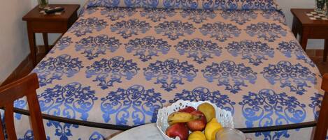 Una cama bien tendida con una colcha azul estampada con flores, una mesita con fruta y una jarra de jugo, y dos lámparas fijadas a la pared.