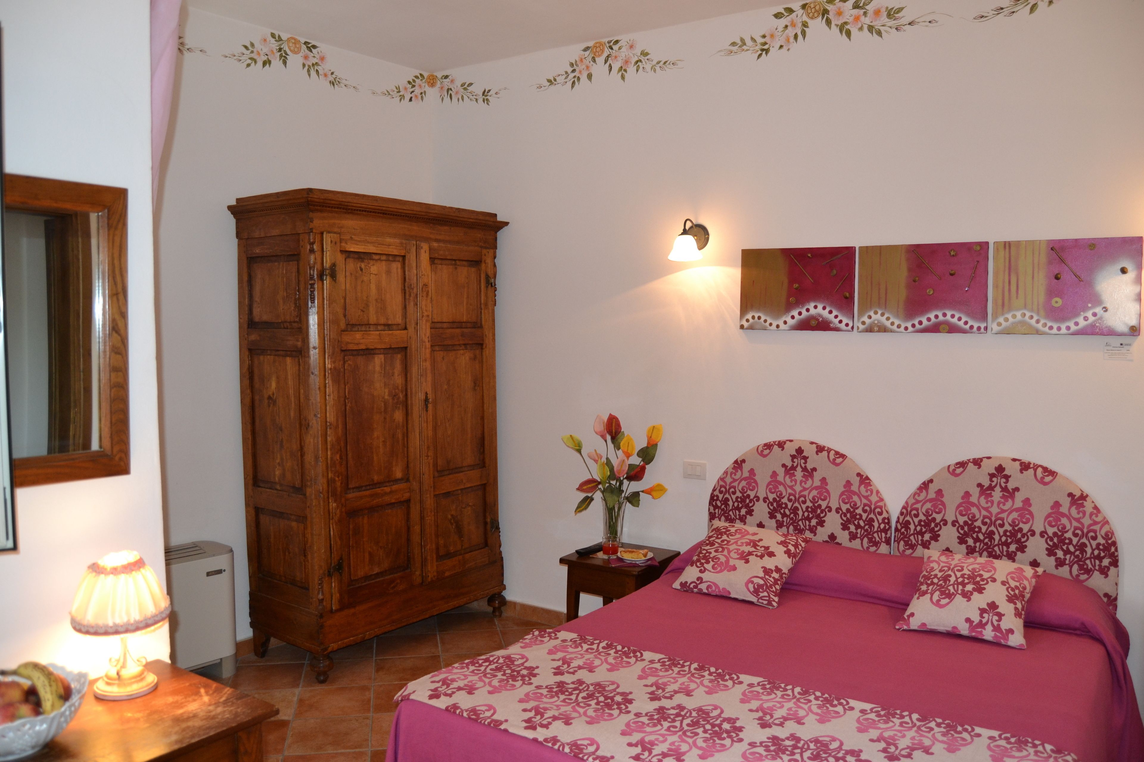 A bedroom with a bed, wooden wardrobe, wall-mounted artwork, and a bedside table with a lamp.