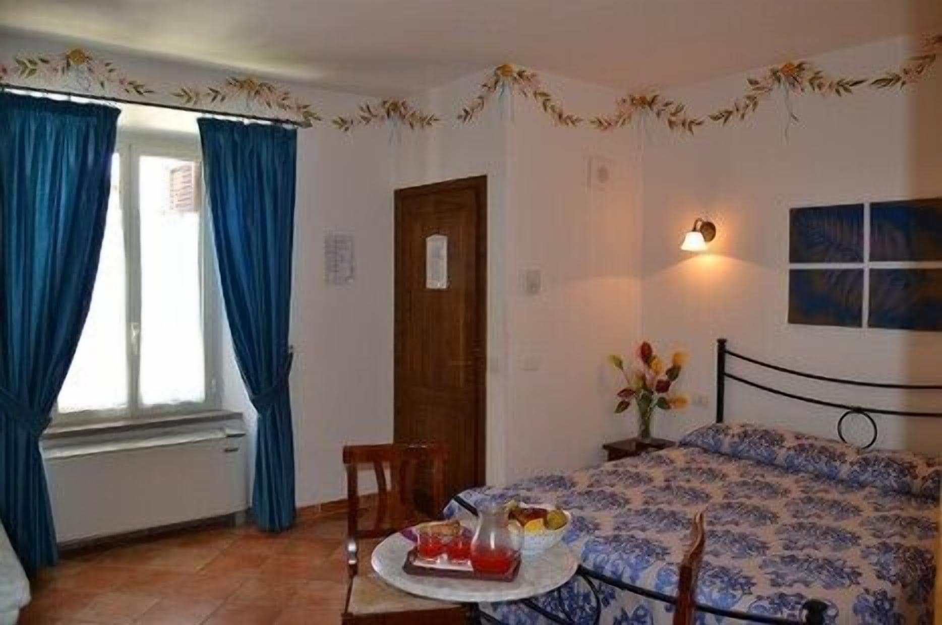 A bedroom with a bed, a chair, a table with a plate and a pitcher, a window with curtains, and a wall-mounted light fixture.