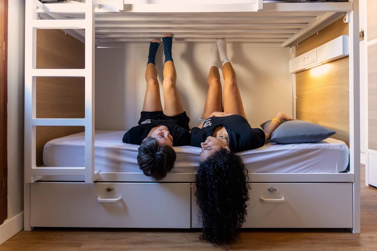 A bunk bed with two people lying on it, a white drawer below, and a wooden panel on the wall.
