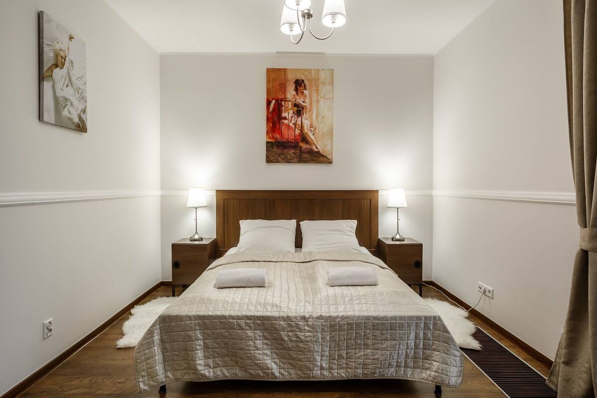 Une chambre à coucher bien aménagée, avec un grand lit, deux tables de chevet avec des lampes, un tableau au mur et un tapis en fausse fourrure blanche.