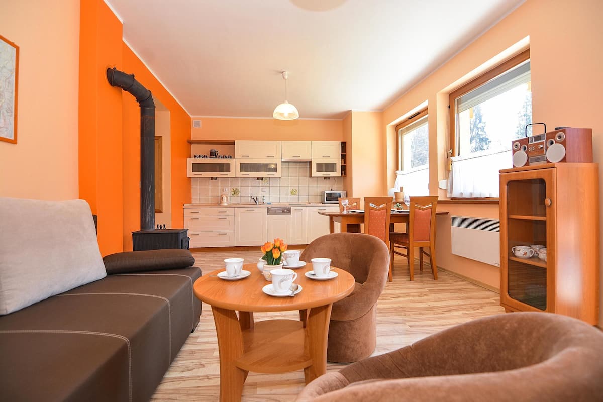 A modern living room with a sofa, a dining table with cups and a vase of flowers, and a kitchen in the background.