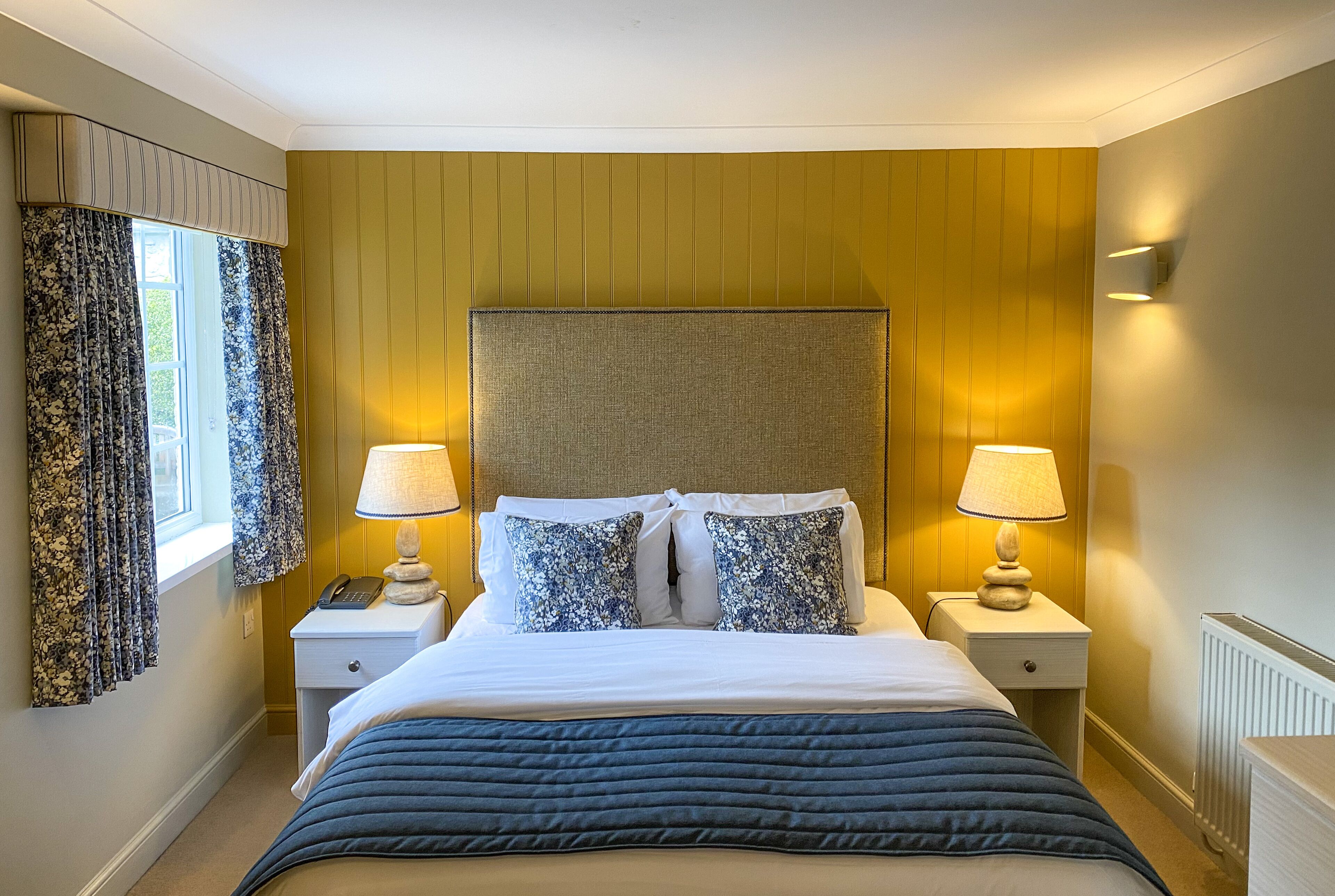 Habitación de hotel con una cama grande, dos mesitas de noche, una lámpara, un teléfono y una ventana con cortinas.