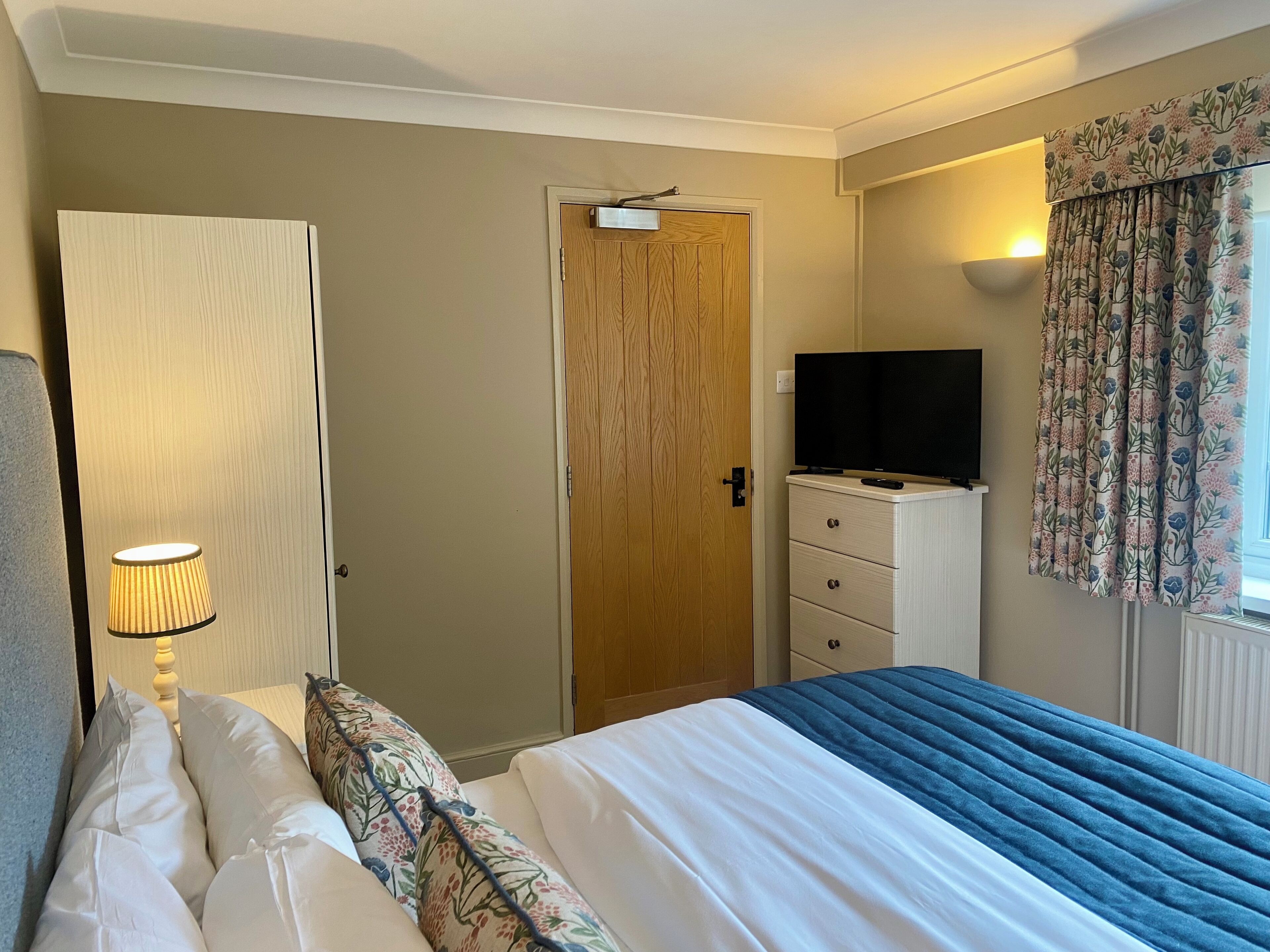 A bedroom with a bed, a dresser, a television, a door, and a window with curtains.
