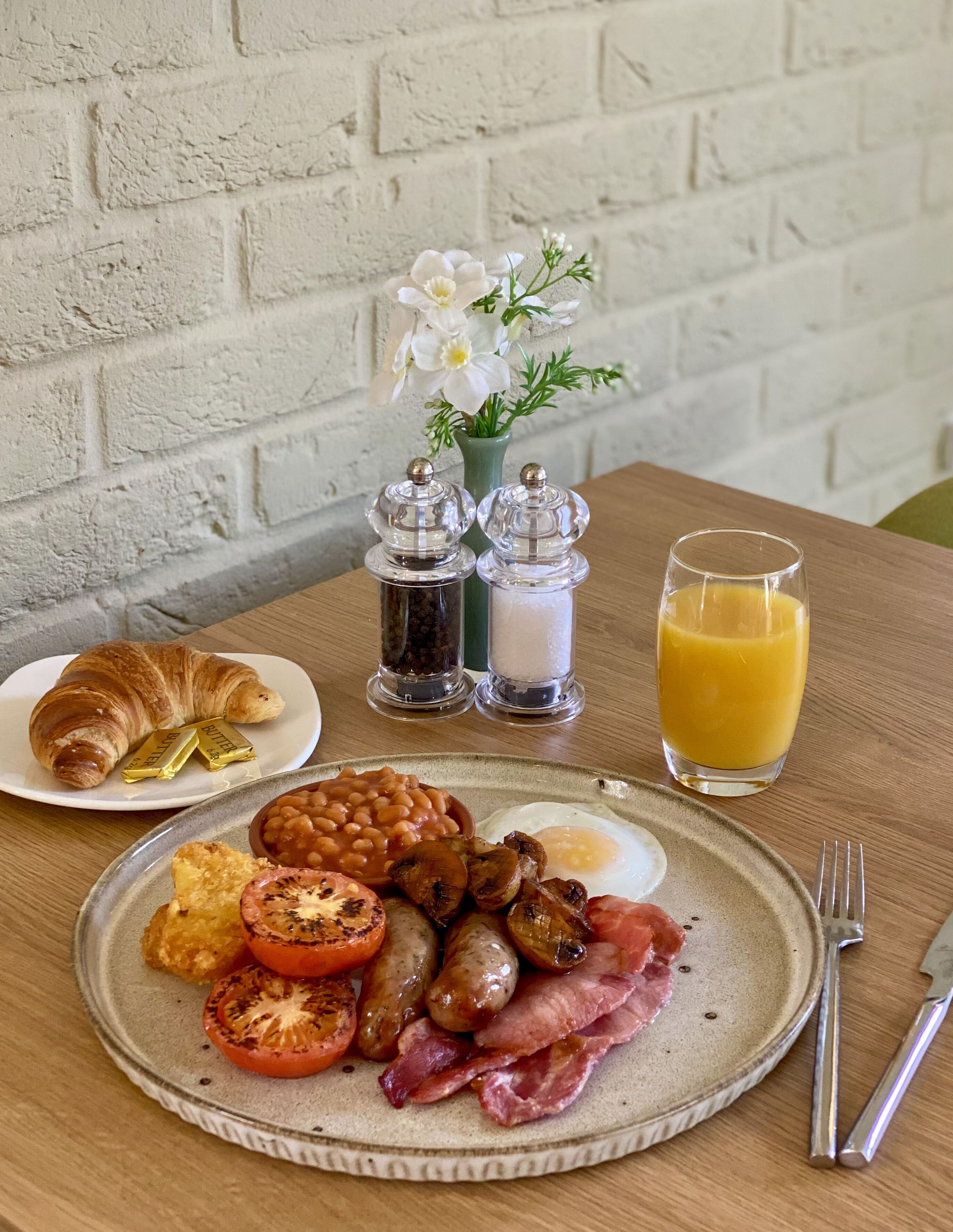 A breakfast plate with eggs, bacon, tomatoes, beans, sausages, and a croissant, along with a glass of orange juice and condiments on a table.