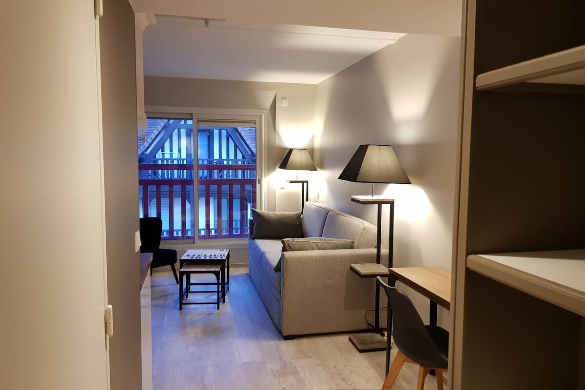 Una sala de estar moderna con un sofá, una mesa de centro y dos lámparas. Unos puertas corredizas de vidrio dan a un balcón con barandilla roja.