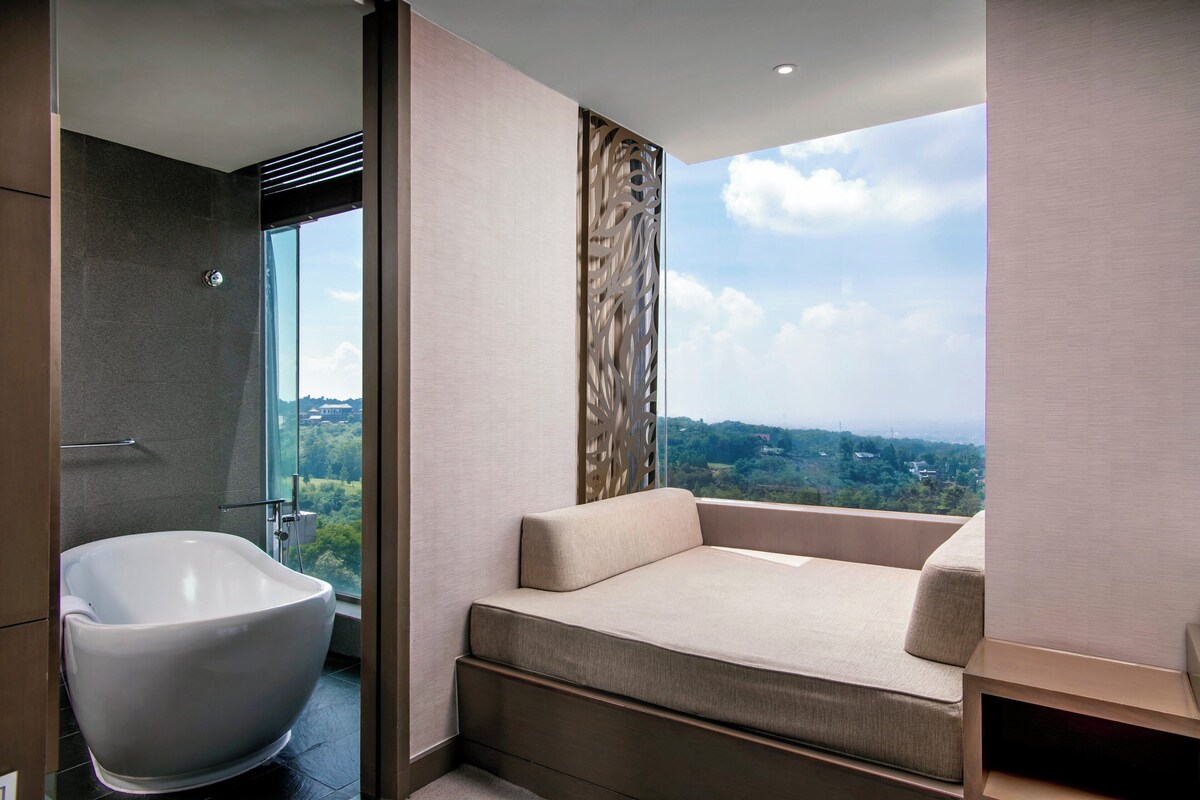 A modern bathroom with a freestanding bathtub, a sofa bed, and a large window offering a city view.
