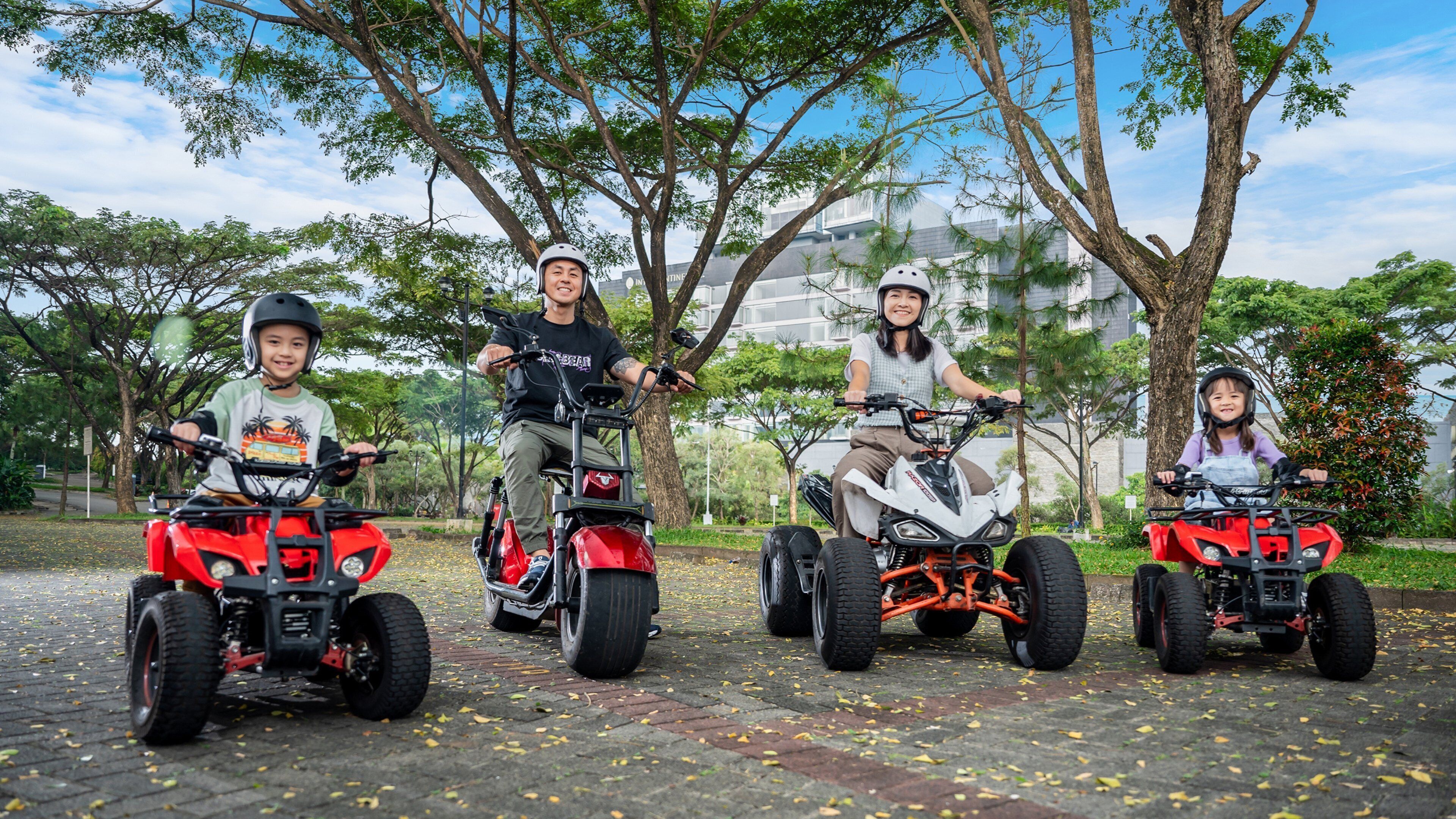 Four people riding ATVs on a paved path surrounded by trees and buildings.