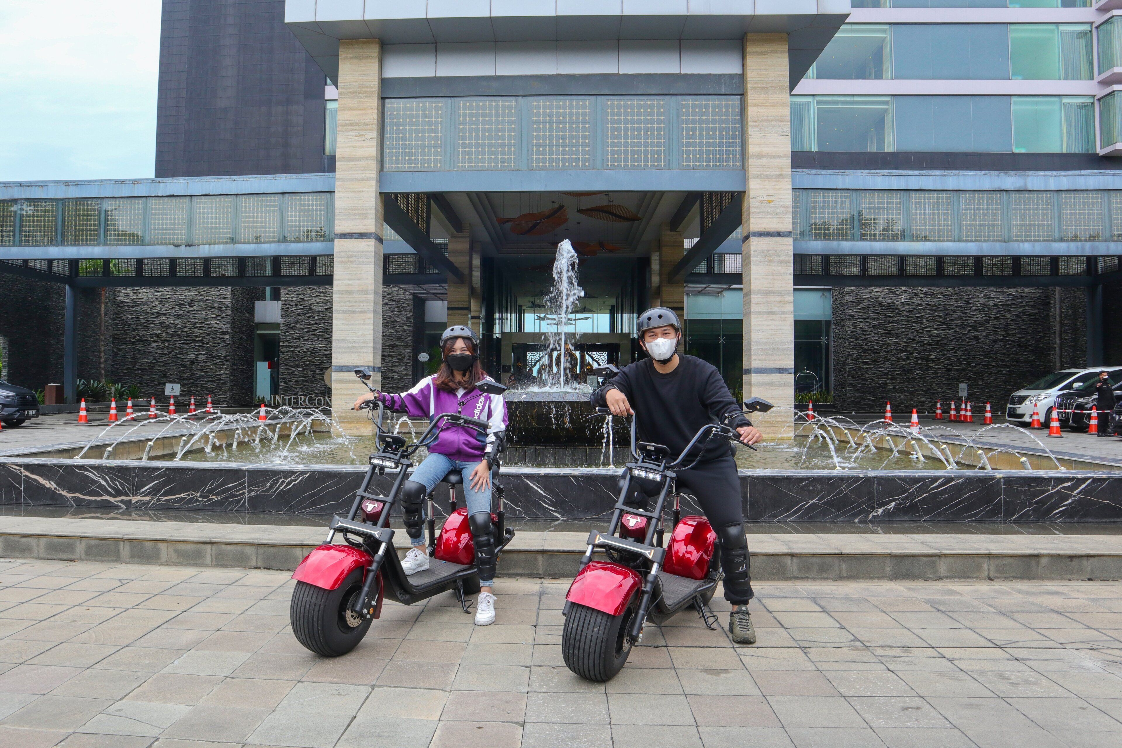 Two people on electric scooters in front of a building with a fountain.