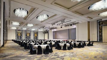 A large banquet hall with rows of tables set for a formal event, featuring chandeliers and a carpeted floor.