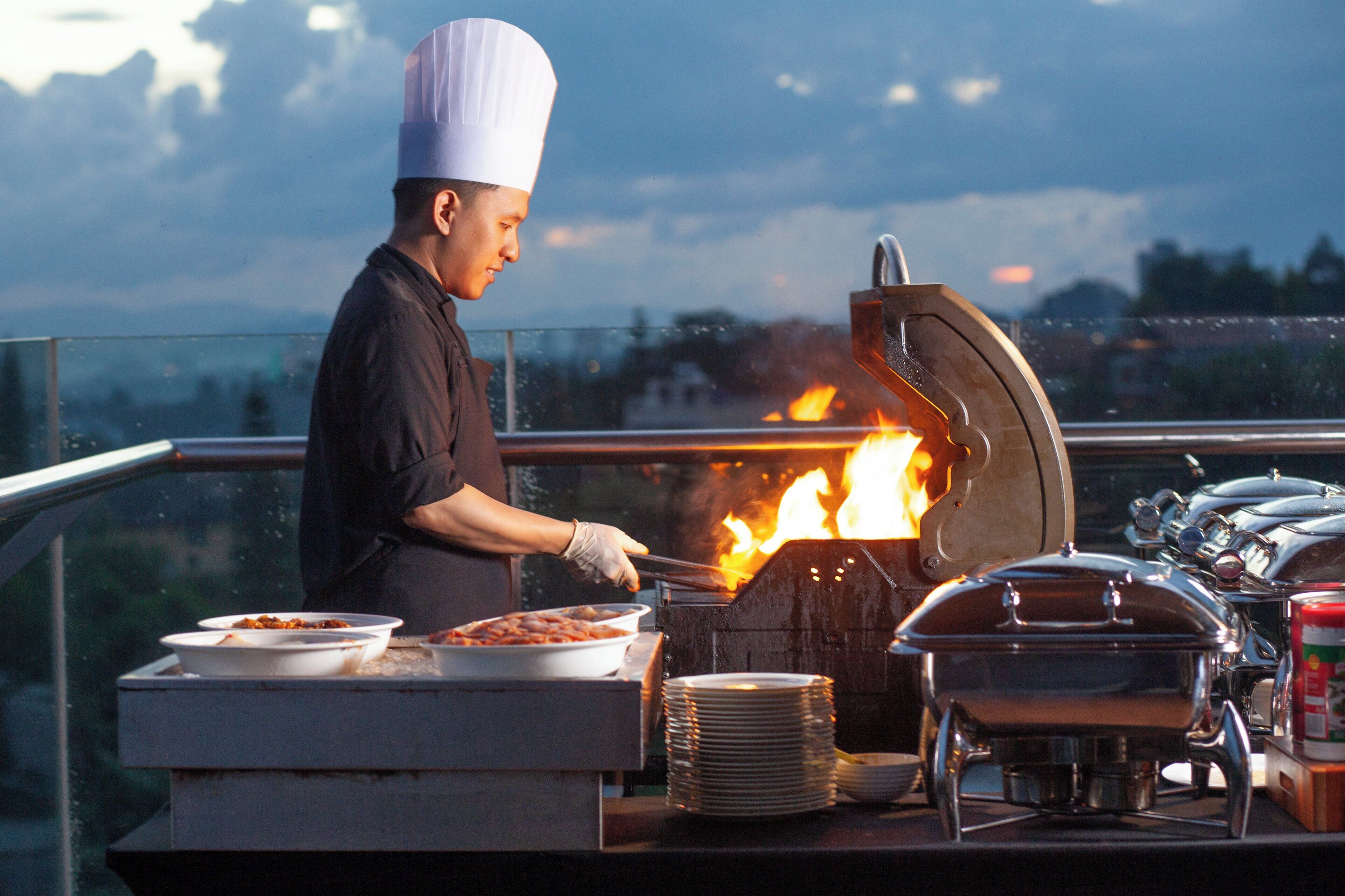 A chef preparing food on an outdoor grill with a cityscape in the background.
