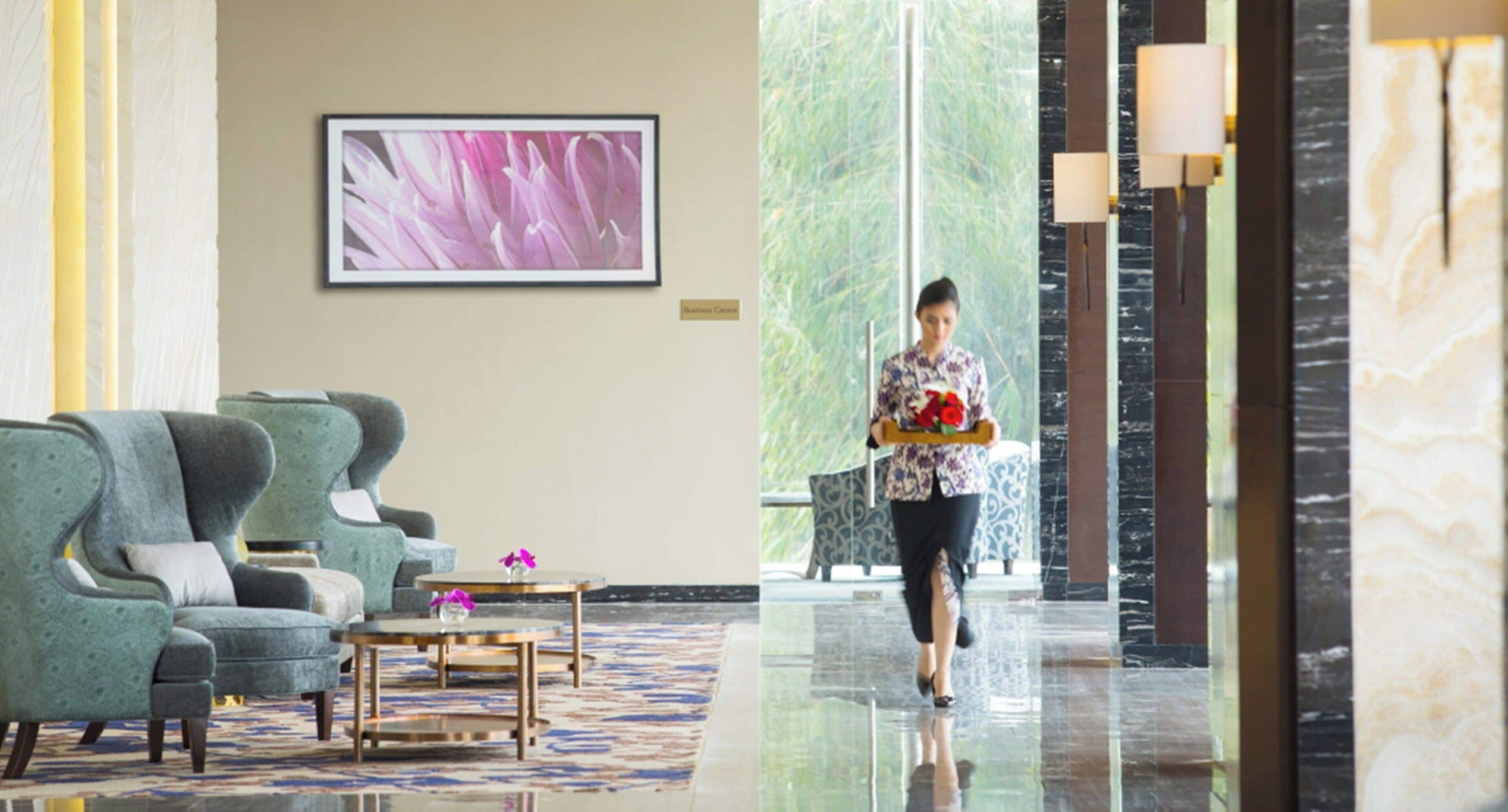 A hotel lobby with seating, a framed floral artwork, and a person carrying a tray with flowers.