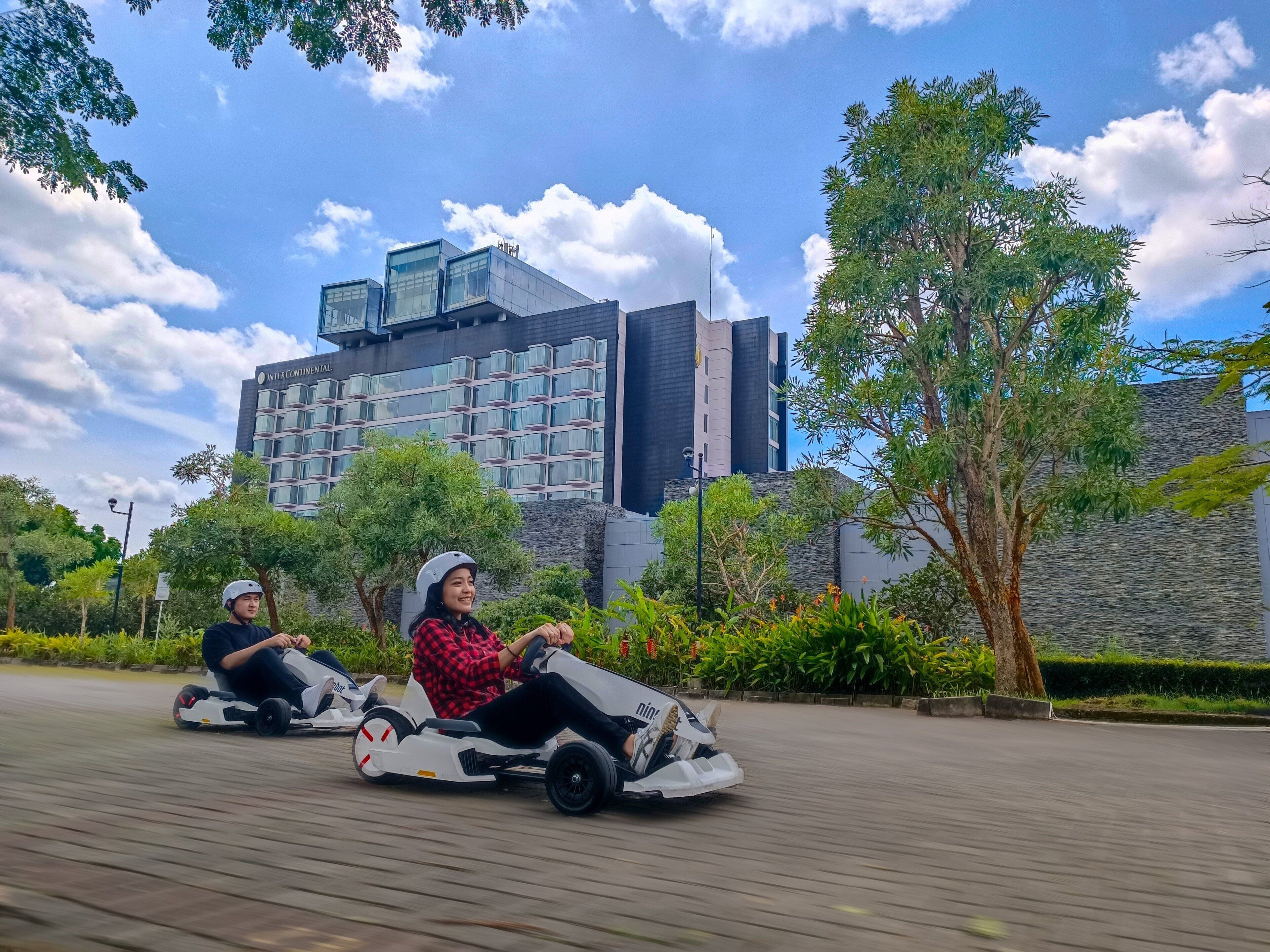 Two people on go-karts on a paved path with a modern building in the background.