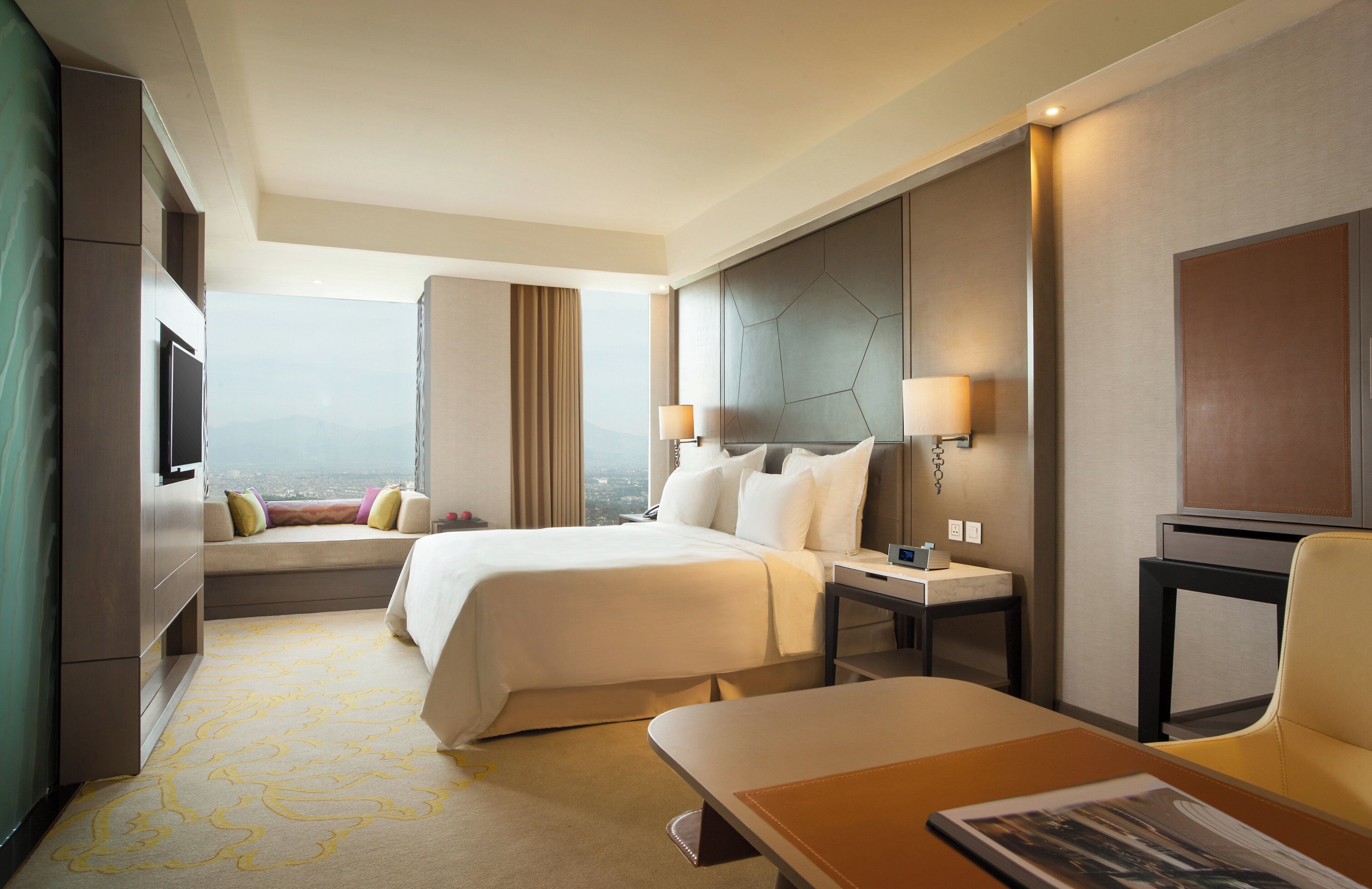 A hotel room with a large bed, a desk, a chair, a TV, and a view of the city.