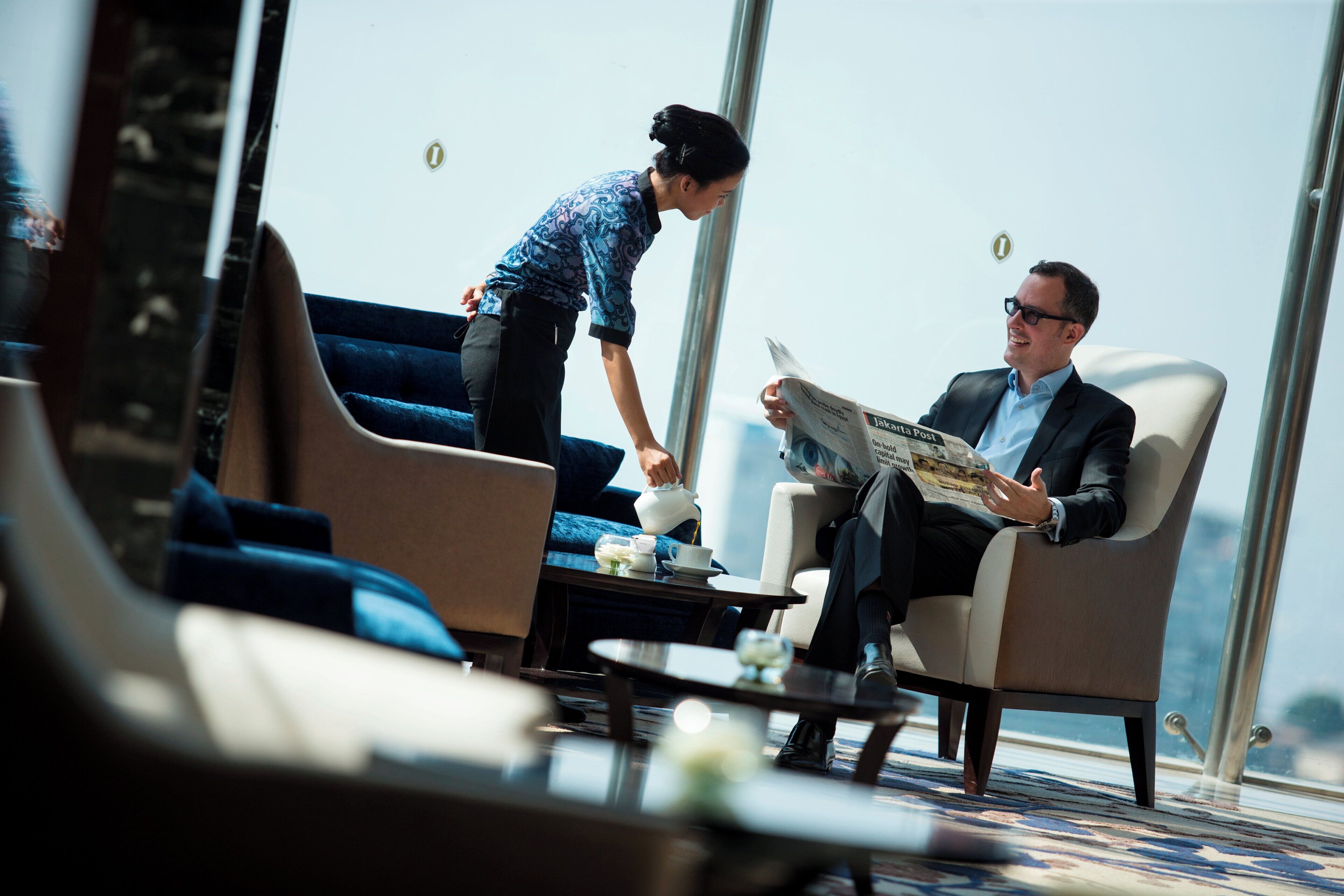 Un homme est assis sur une chaise et lit un journal, une autre personne se tient à côté de lui, et une table basse est placée à proximité, avec une tasse et une soucoupe dessus.