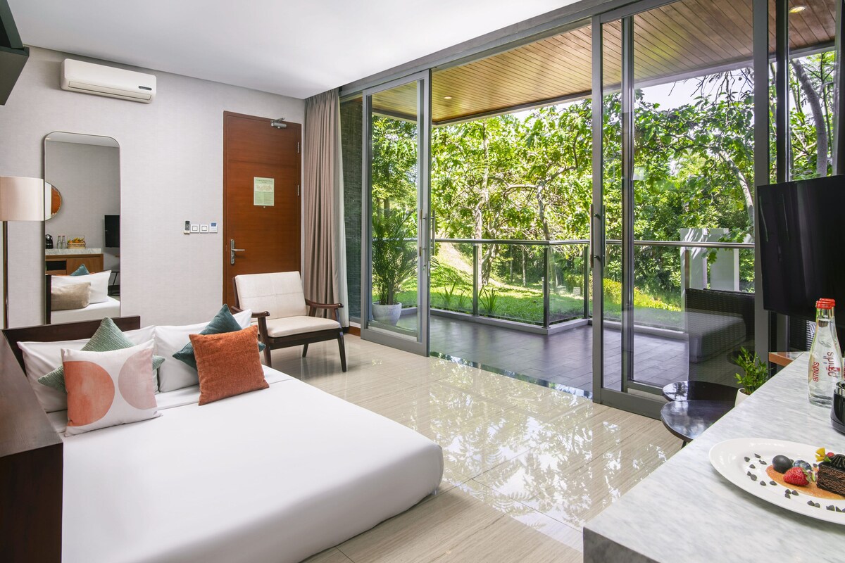 A modern hotel room with a large sliding glass door leading to a balcony, a white sofa with decorative pillows, a small table with a bottle and a plate of food, and a mirror on the wall.