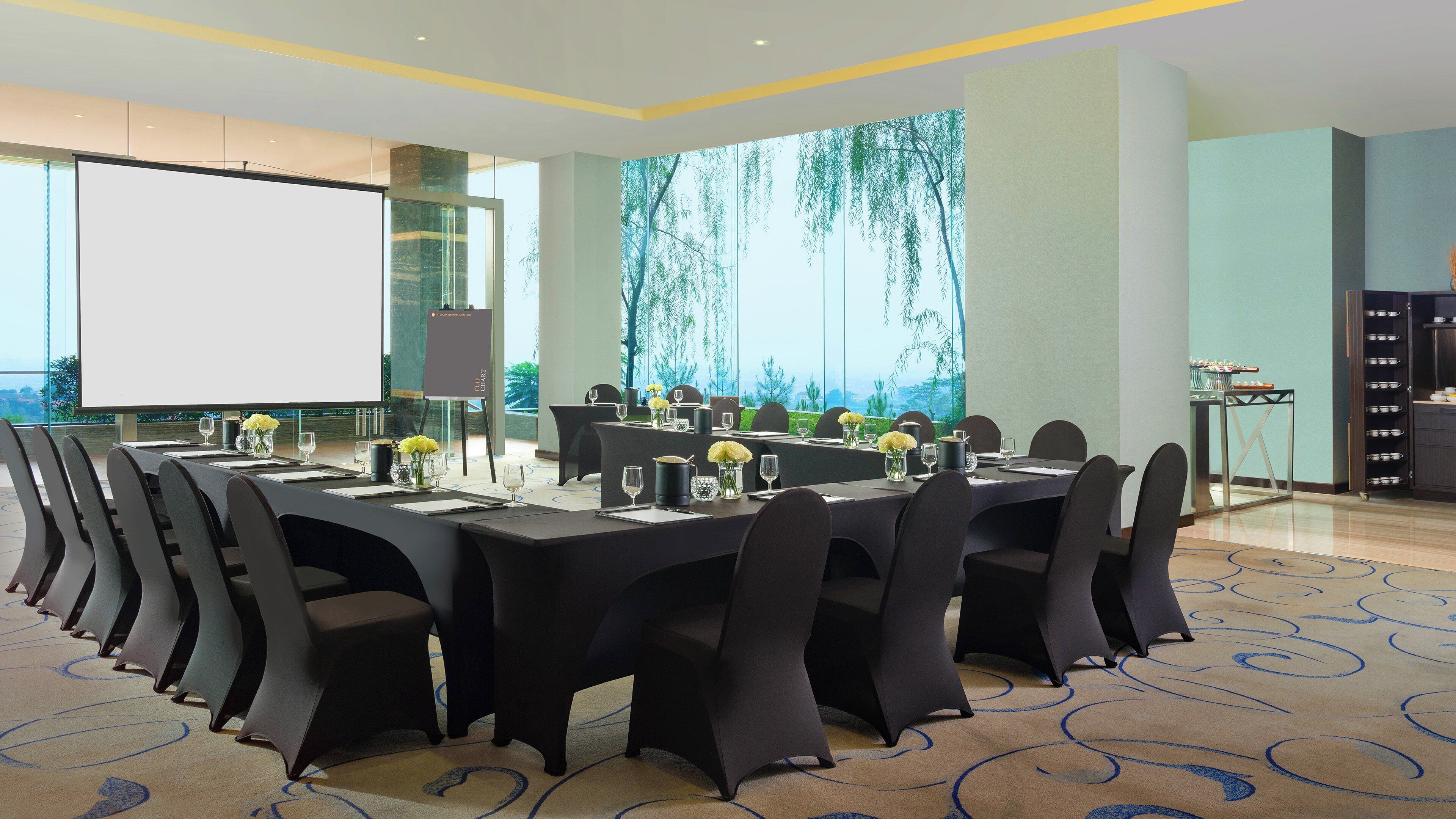 A conference room with a large screen, multiple tables set for dining, and a view of trees through the windows.