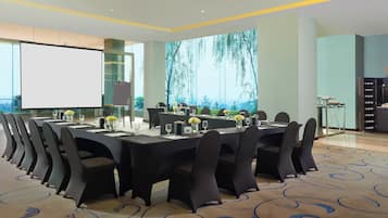 A conference room with a large screen, multiple tables set for dining, and a view of trees through the windows.
