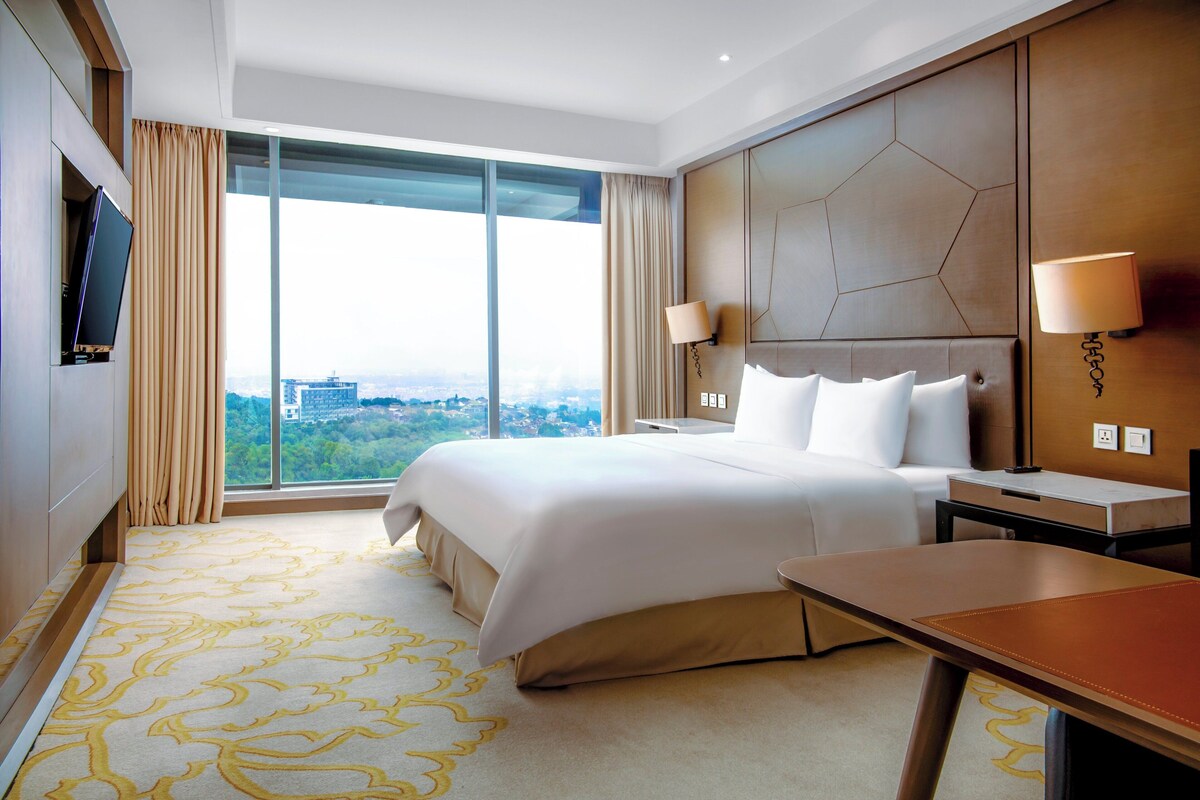 A hotel room with a large bed, a desk, a TV, and a view of the city.