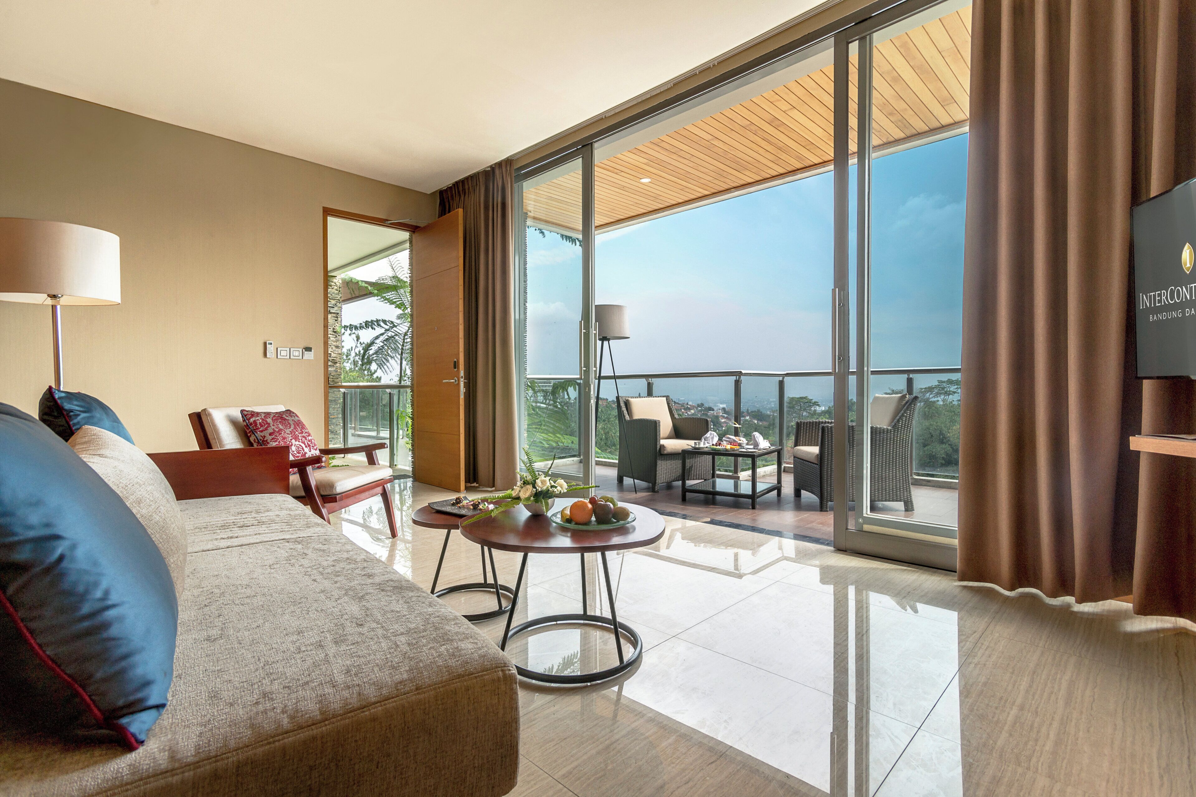 A modern hotel room with a large sliding glass door leading to a balcony, a sofa, a small table with a vase of flowers, and a television on the wall.