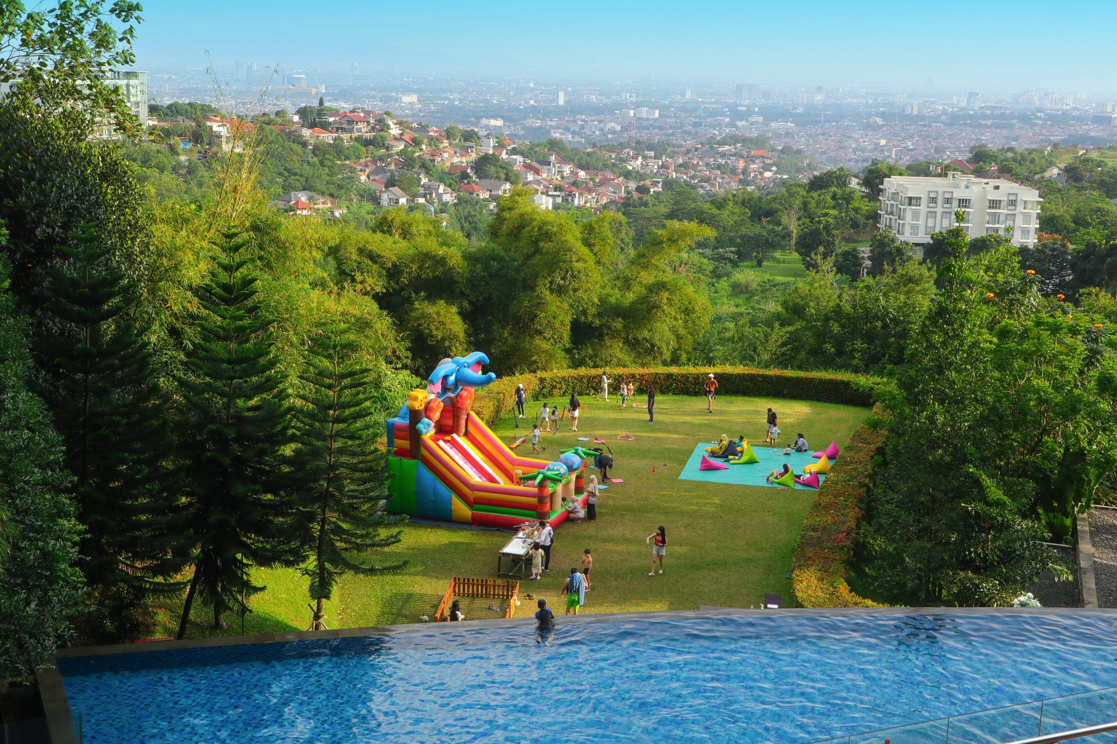 A swimming pool with a slide and inflatable play area, surrounded by trees and a view of a cityscape.