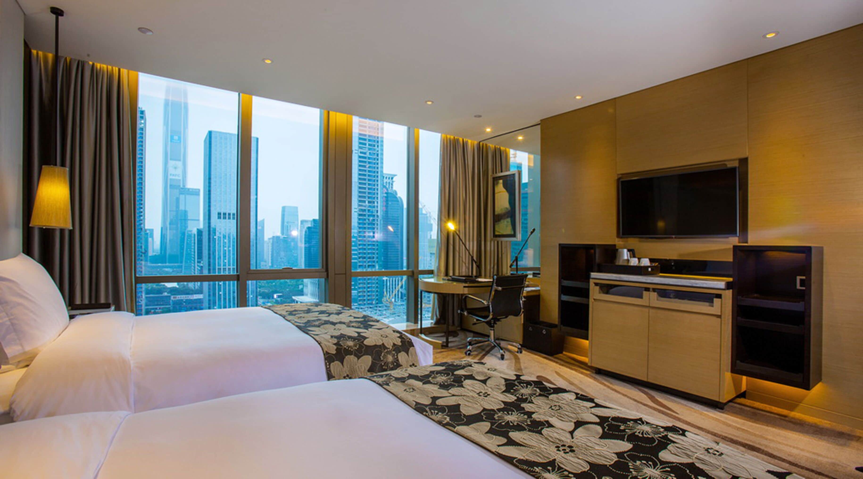 A hotel room with two beds, a desk, a TV, and a city view.