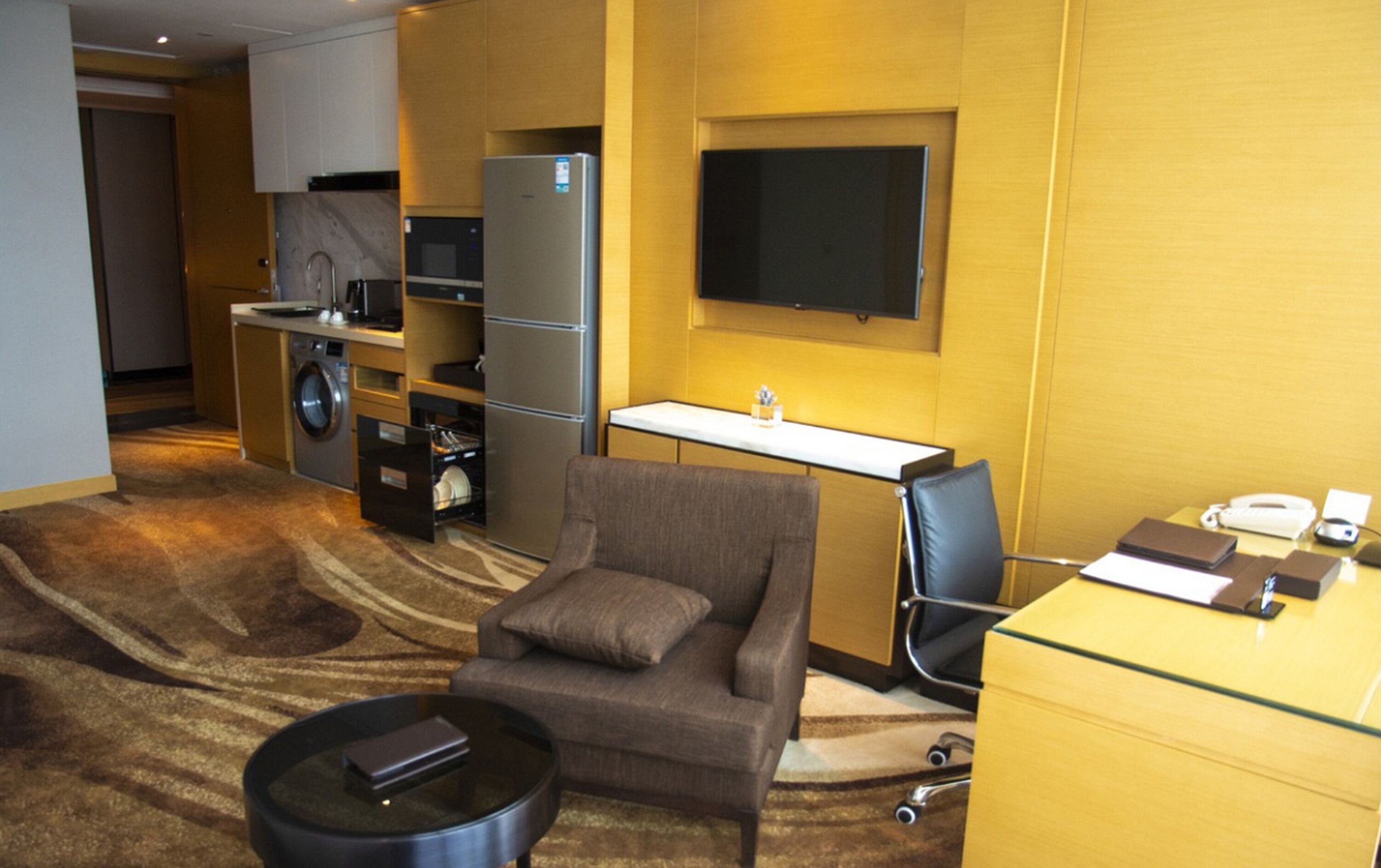 A compact hotel room with a sofa, a small coffee table, a desk with a computer, a washing machine, and a refrigerator.