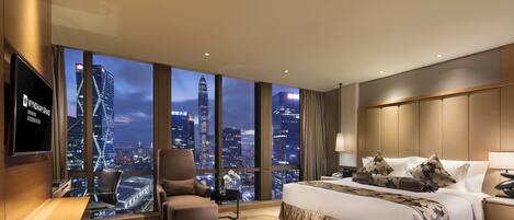 A hotel room with a large bed, a desk, a chair, and a TV. The room has a view of a city skyline at night.