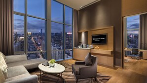 Una habitación de hotel moderna con una amplia vista de la ciudad, televisor de pantalla plana, sofá, una silla y una pequeña mesa con una planta.