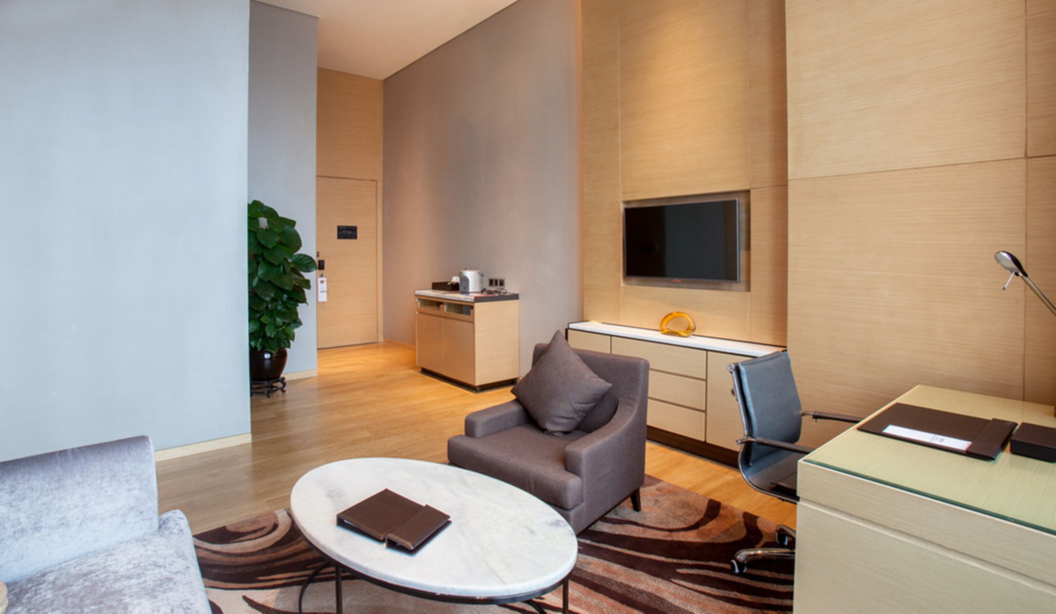 Una habitación de hotel moderna con un sofá, una mesita redonda pequeña, un escritorio con una computadora, un televisor y una planta en maceta.