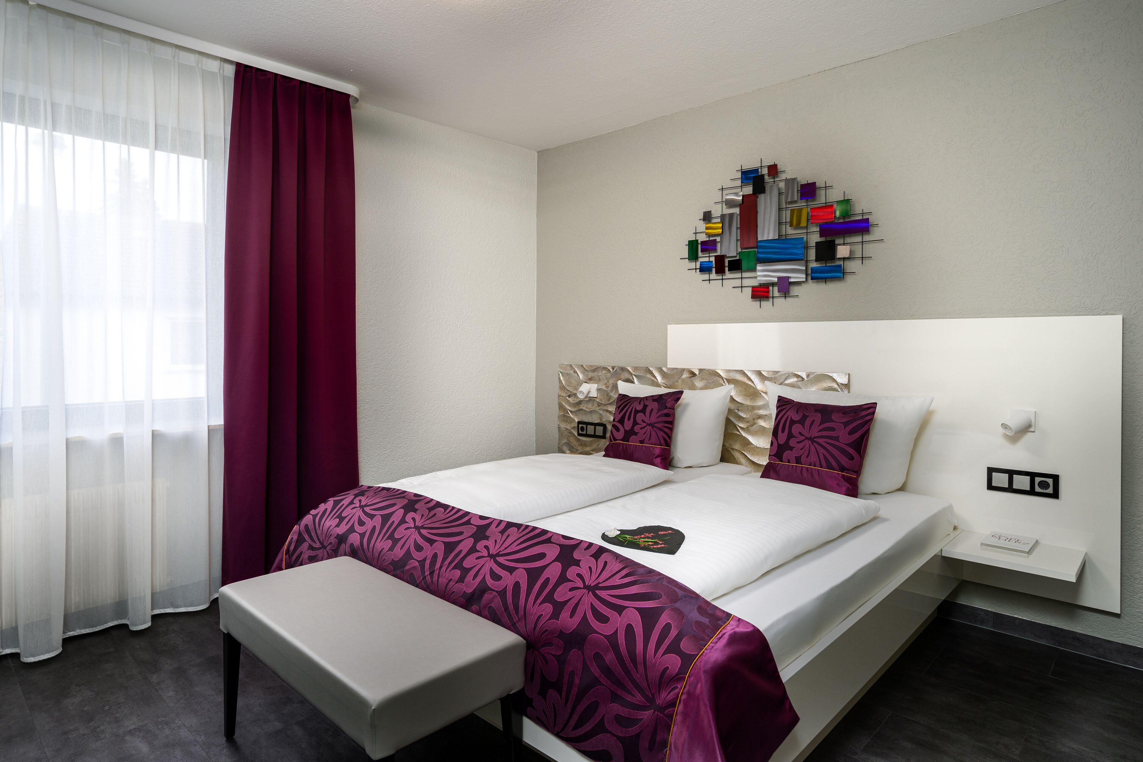 Una habitación de hotel con una cama grande, una mesita de noche, un adorno en la pared y una ventana con cortinas traslúcidas.