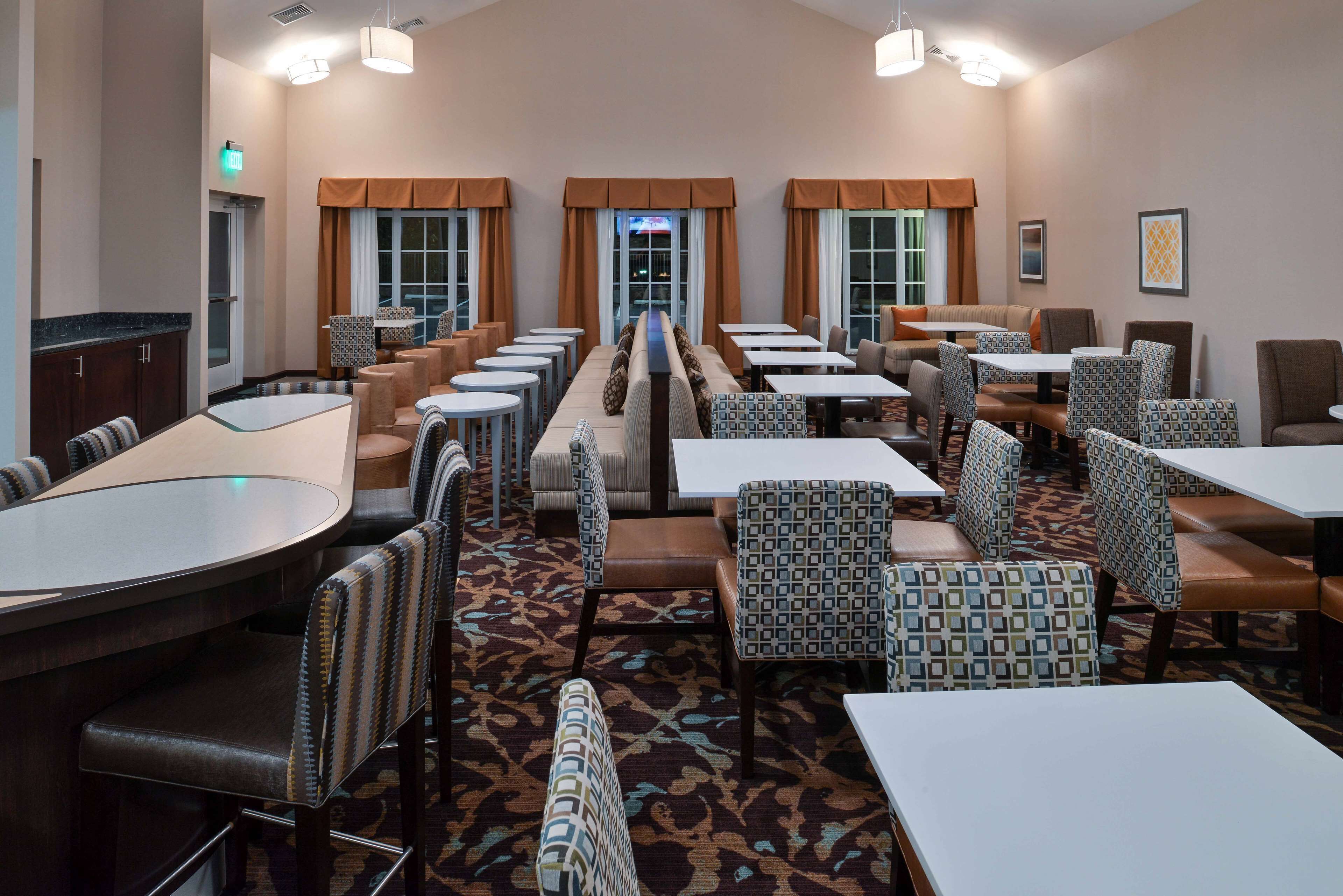 Een restaurantgedeelte in een hotel met meerdere tafels en stoelen, een kamerbreed tapijt en uitzicht door de ramen.