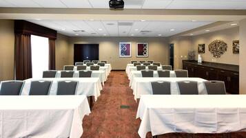 Un salón de conferencias con filas de mesas y sillas, un proyector y un ventanal con cortinas.
