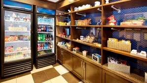 A self-service breakfast area with refrigerated beverage dispensers, shelves stocked with snacks, and a check-in counter. - Hampton Inn Butler (Butler)