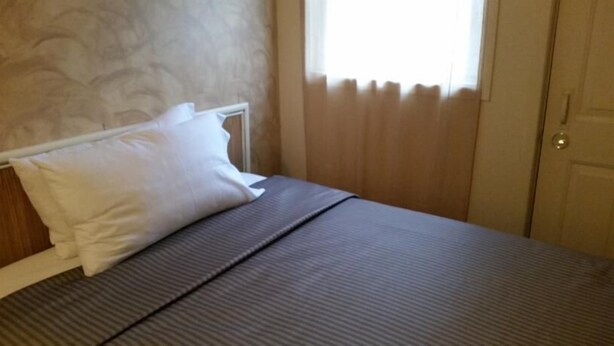 Una cama bien hecha con una almohada blanca y una colcha gris en una habitación con paredes color beige y una ventana con persianas.
