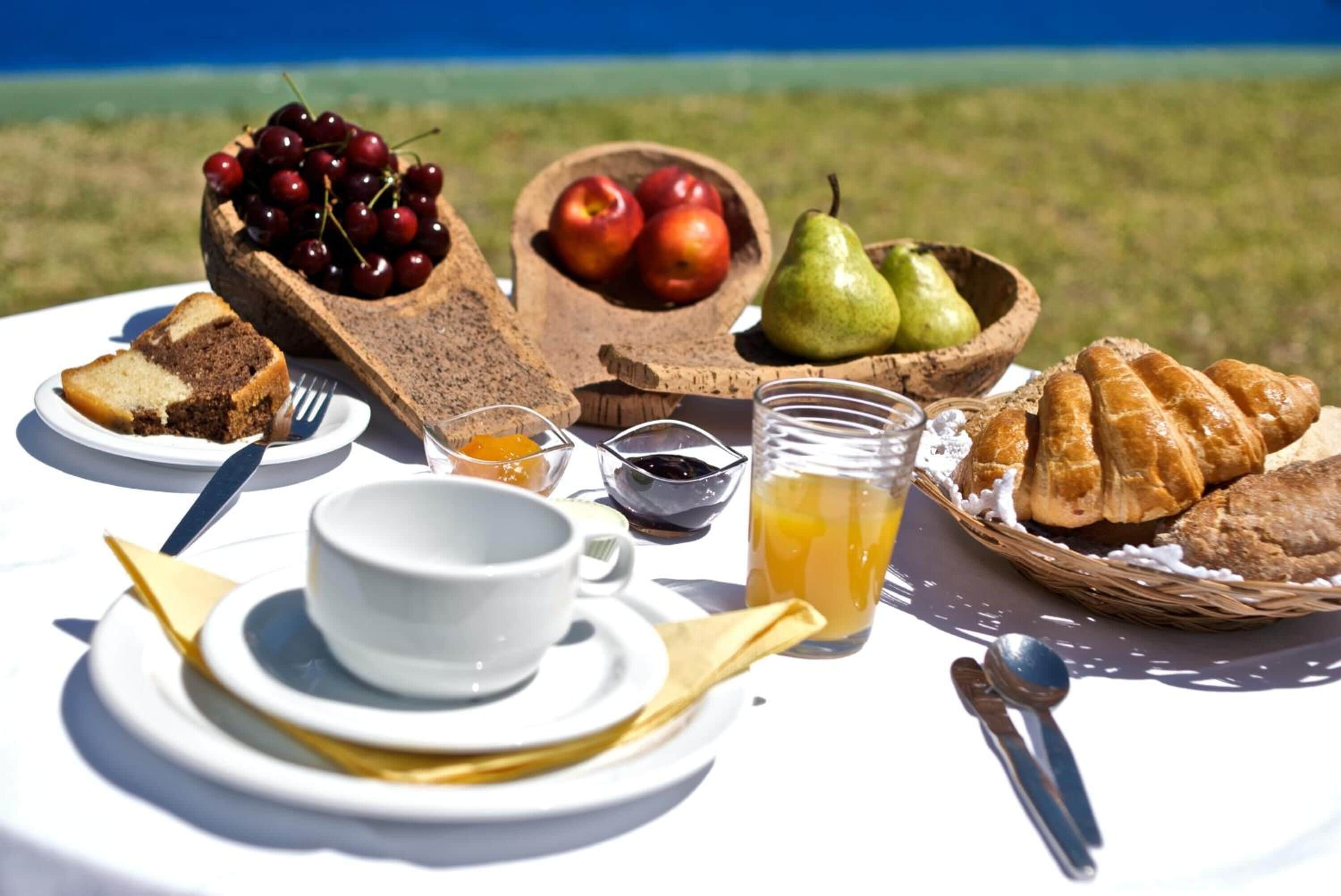 Una mesa dispuesta con una porción de pastel, un cuenco de cerezas, un cuenco de manzanas, una pera, un vaso de jugo de naranja y una cesta de cruasanes.