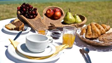 Una mesa dispuesta con una porción de pastel, un cuenco de cerezas, un cuenco de manzanas, una pera, un vaso de jugo de naranja y una cesta de cruasanes.