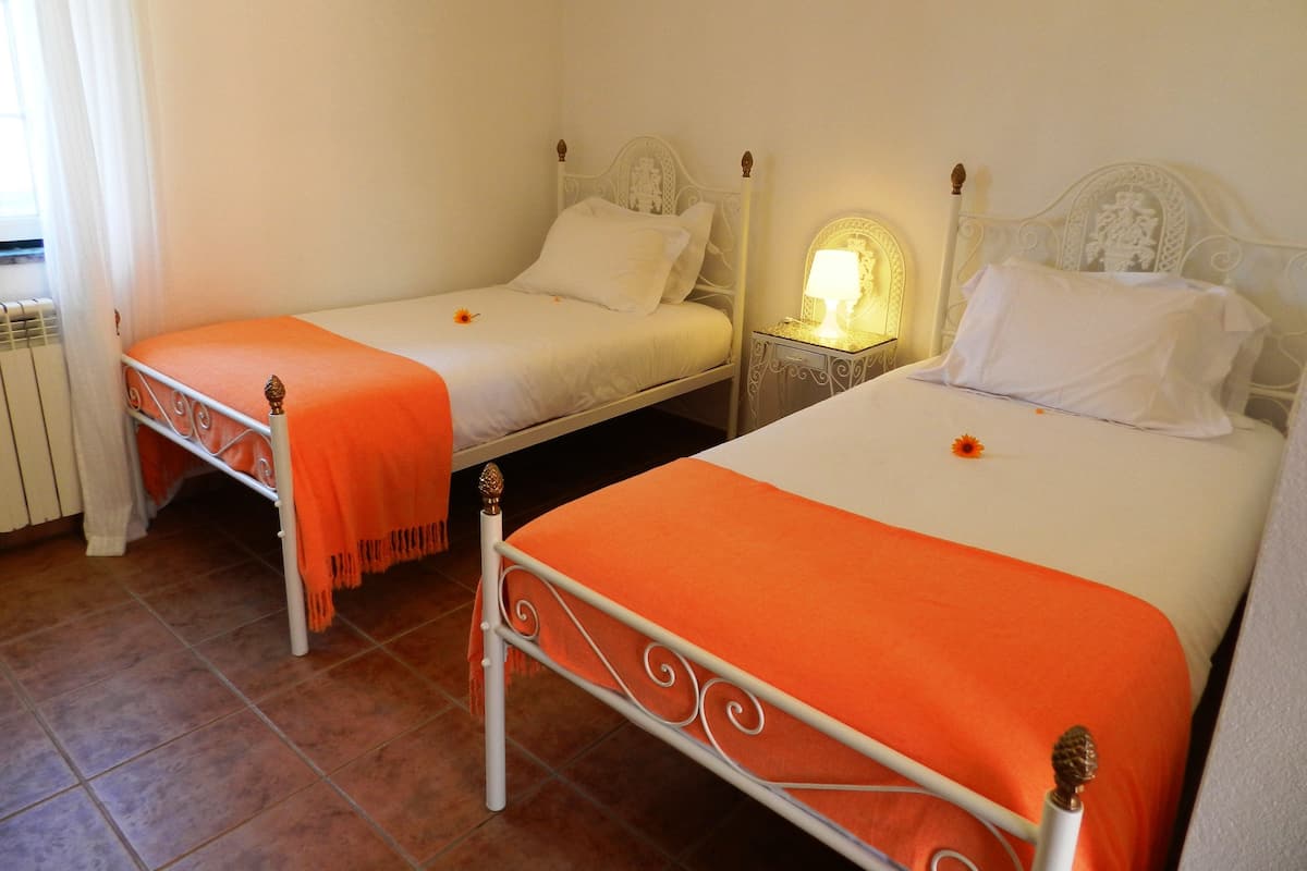 Two single beds with white headboards and orange bedspreads, a nightstand with a lamp, and a window with curtains.