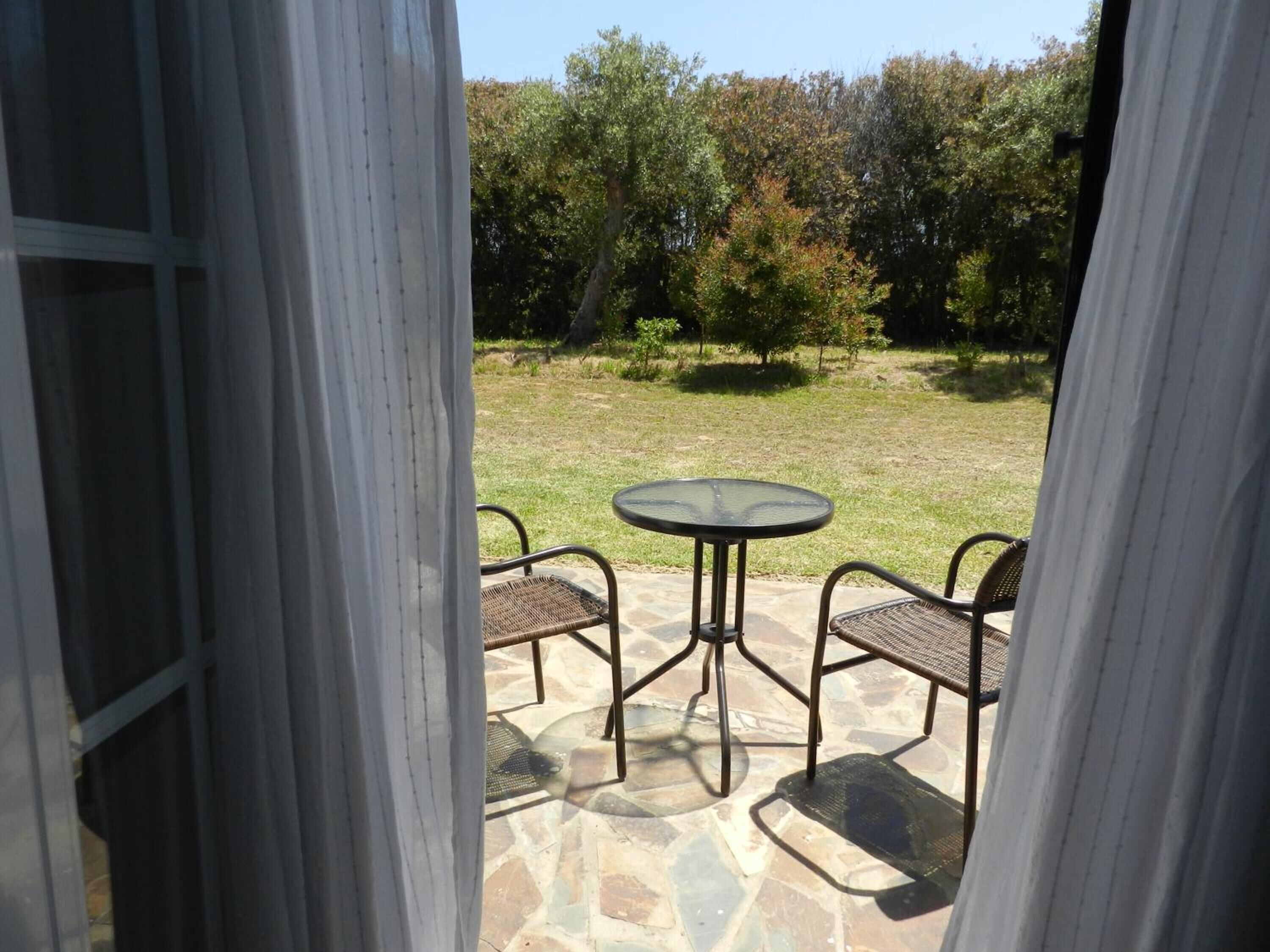 Un patio al aire libre con una mesa redonda y dos sillas, piso de piedra y vistas a un área verde con árboles.