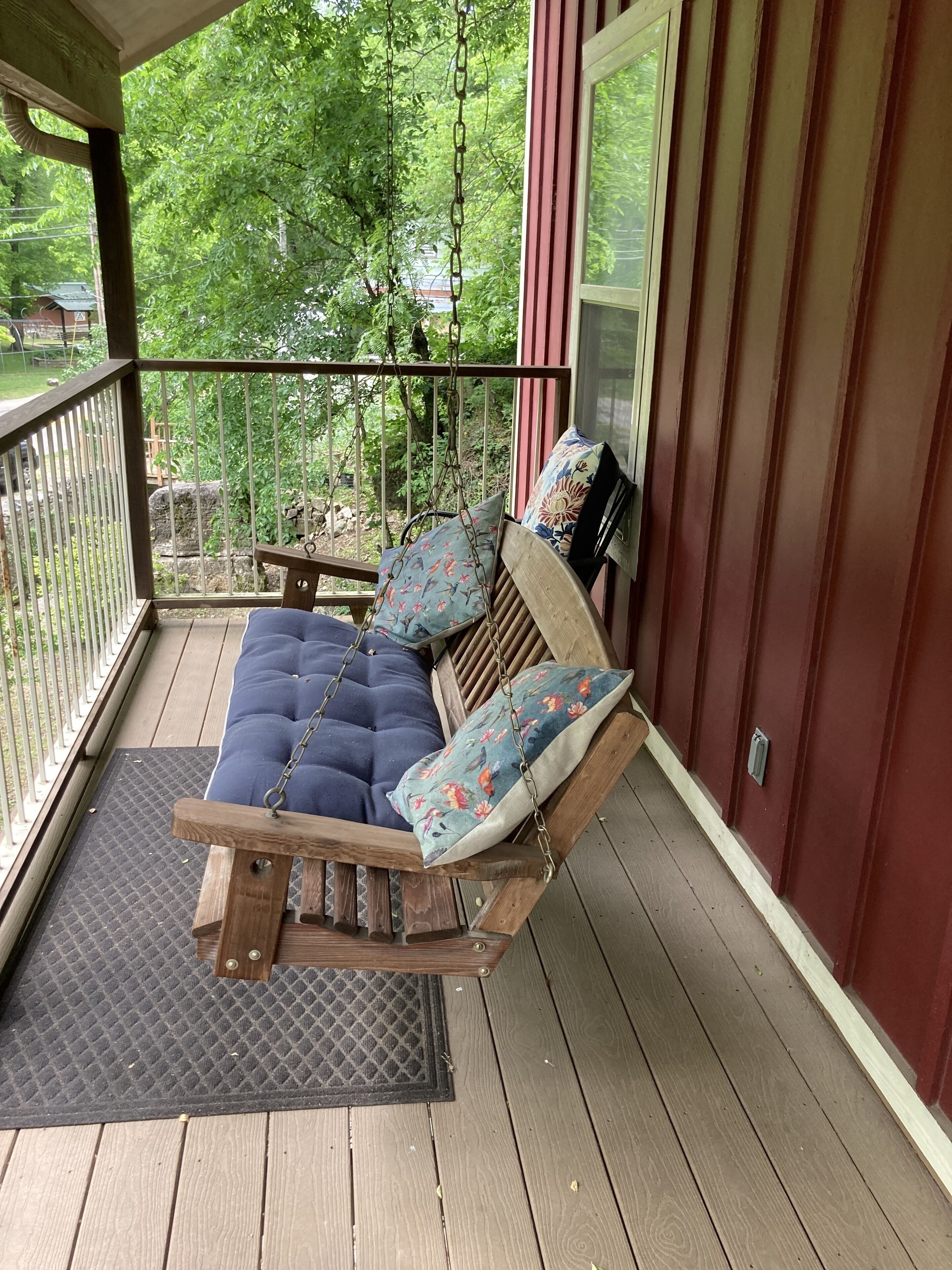 A wooden swing bed with floral cushions on a wooden deck, overlooking a garden.