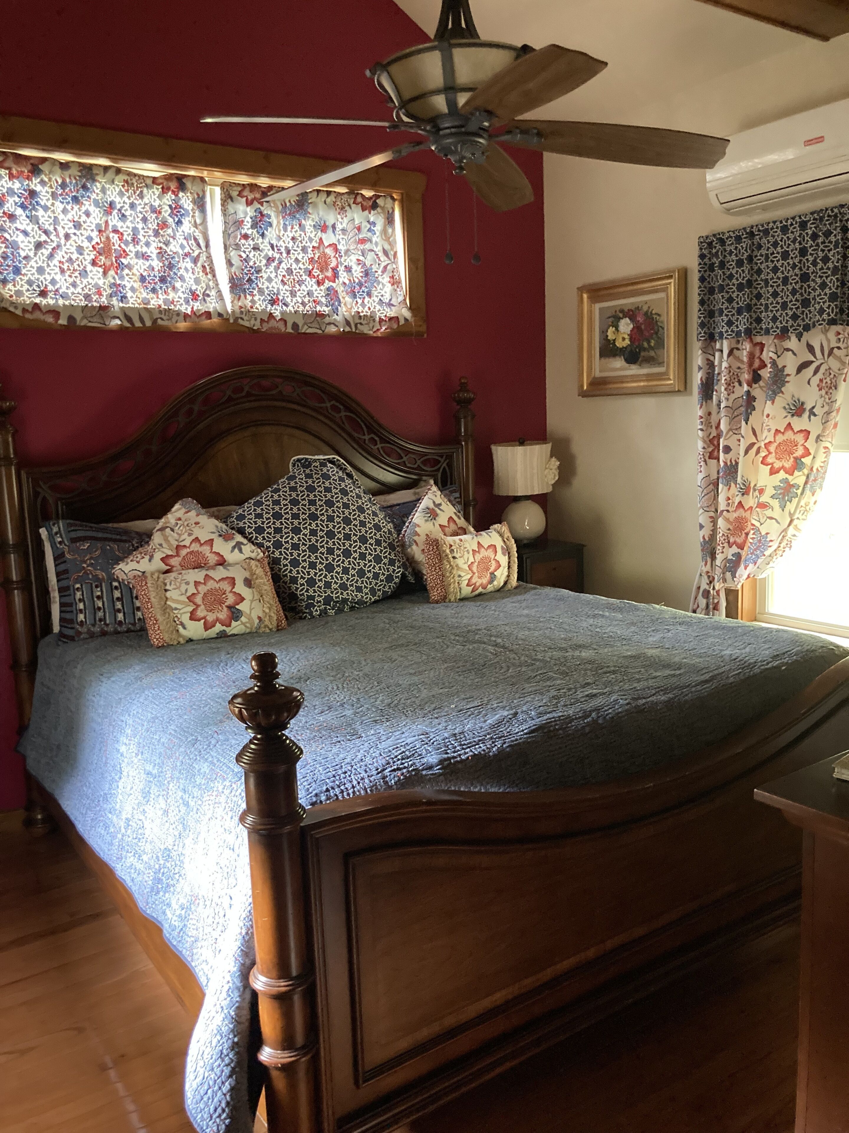 Un dormitorio con cama de madera, cortinas florales, ventilador de techo y ventana con cortinas estampadas.