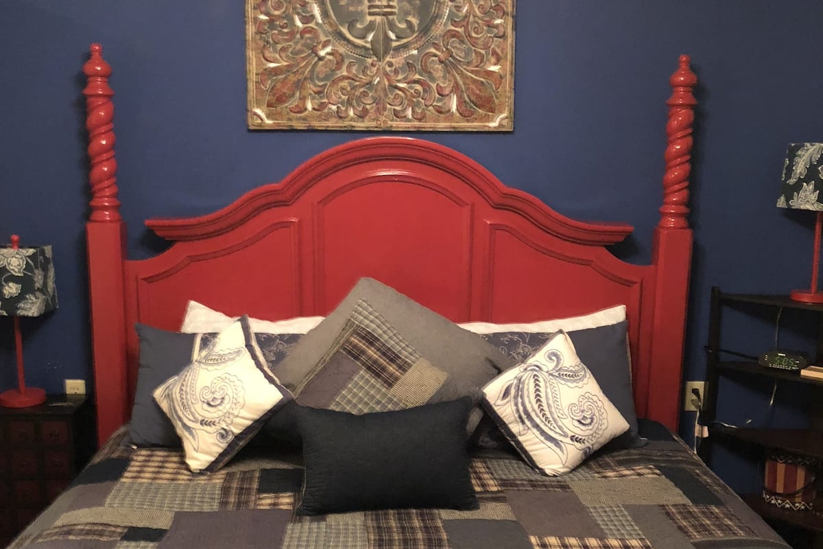 Uma cama com cabeceira vermelha, várias almofadas e colcha estampada.