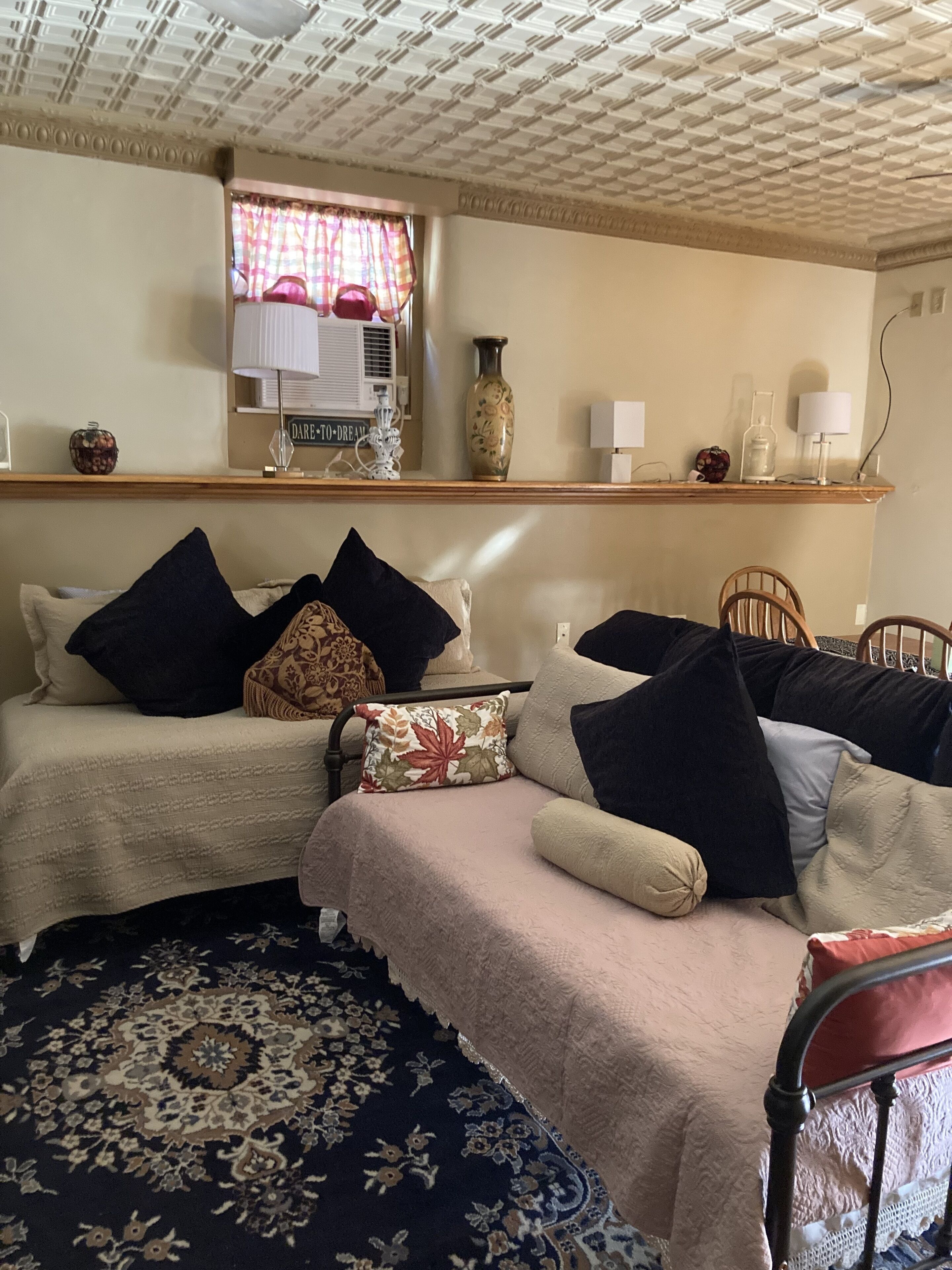 A cozy bedroom with two beds, a wooden shelf with decorative items, a patterned rug, and a window with curtains.