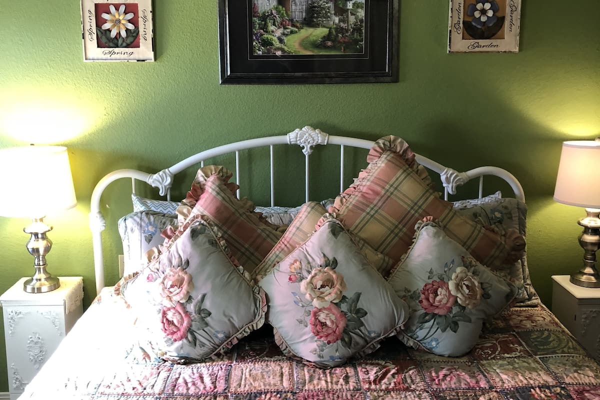 Quarto com parede verde, estrutura de cama de metal branca, roupa de cama florida e xadrez, duas luminárias de cabeceira e quadros com flores e um jardim.