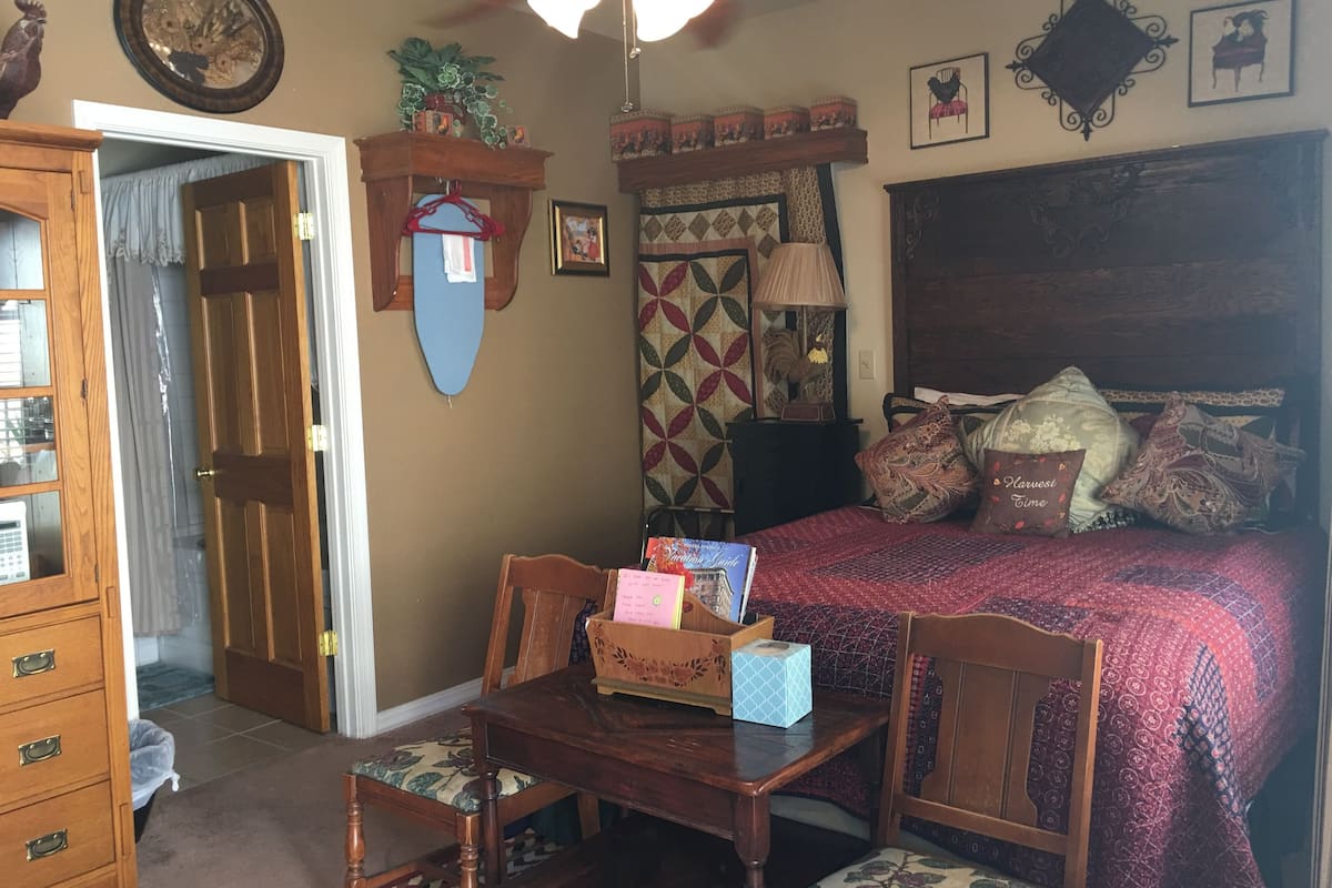 Quarto com cama, escrivaninha e cadeiras. Tem um aparador de madeira e um espelho na parede.