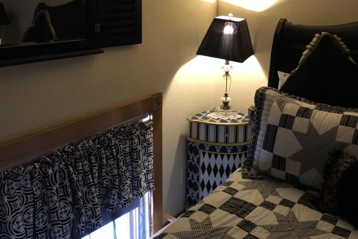 Quarto com cama, luminária de cabeceira, espelho, prateleira com objetos decorativos e janela com cortinas.